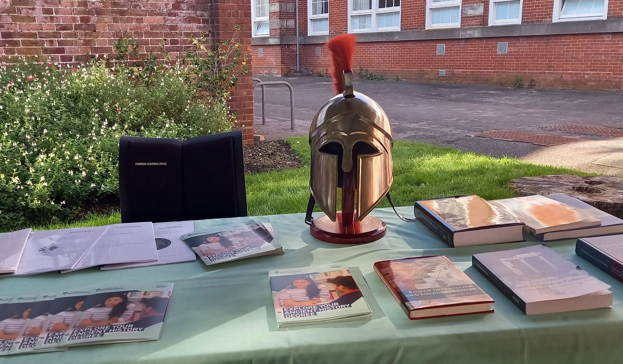 Table set up outdoors with Greek helmet surrounded by brochures and books.