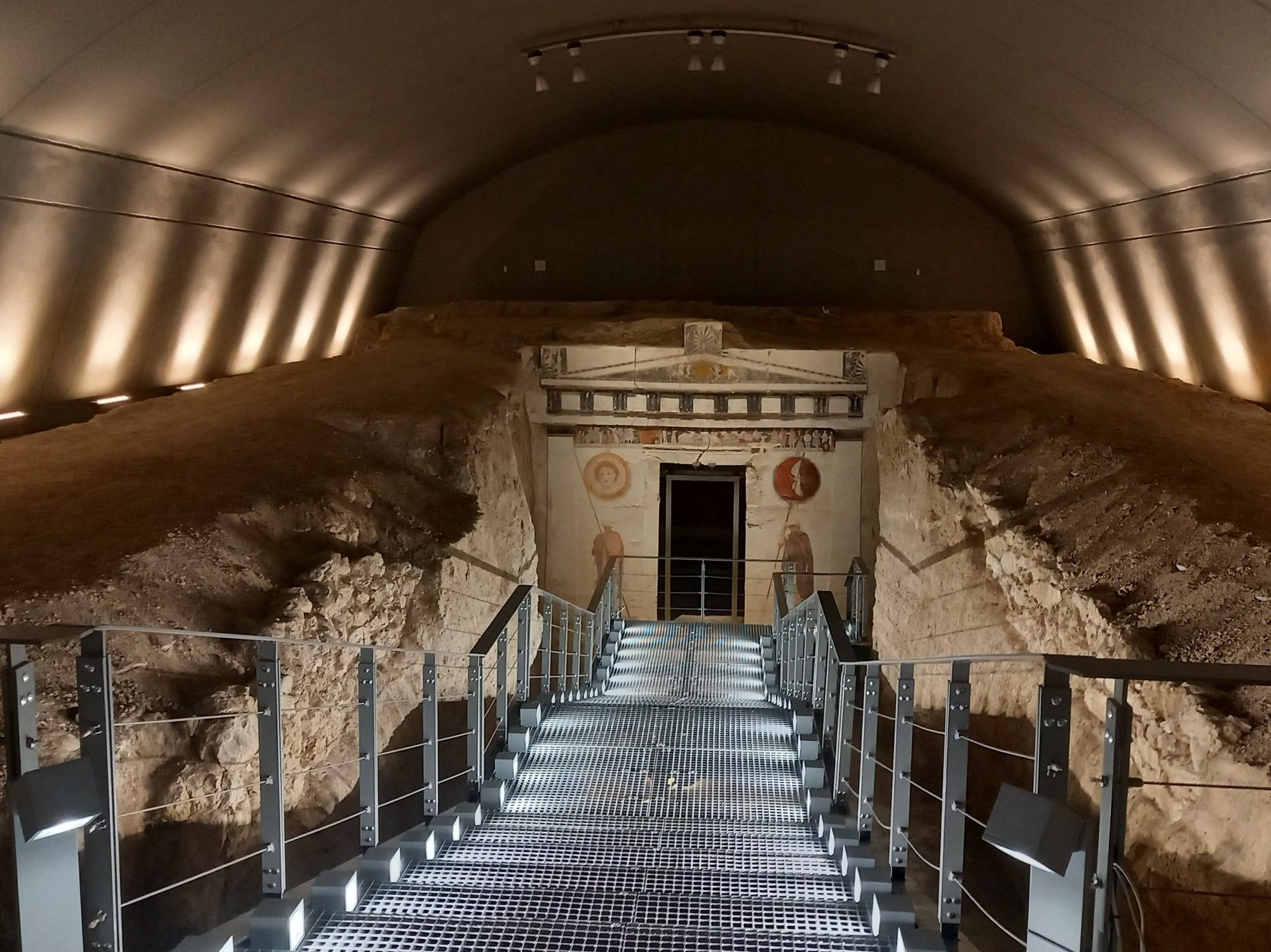 View down a metal staircase leading to the façade of an ancient Macedonian tomb.