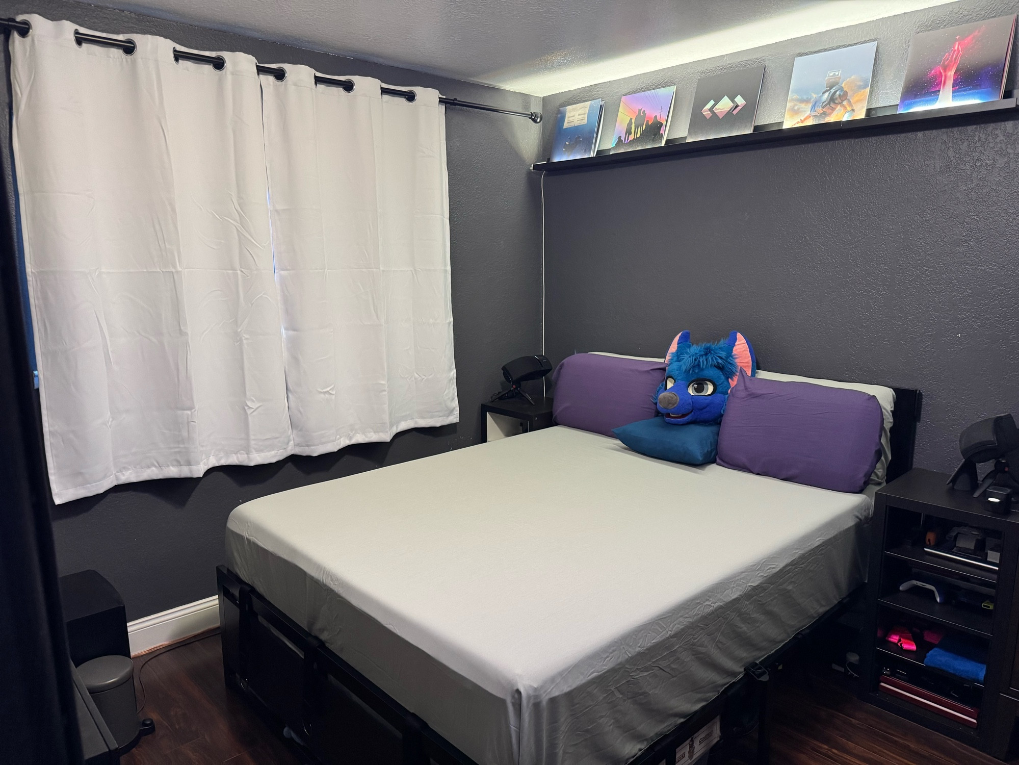 Angled shot of white blackout curtains hanging, bed with white sheets, as well as purple pillows in front of white pillows, in between is one blue pillow with Sybers head sitting on it, 4 vinyl albums sitting above the bed 