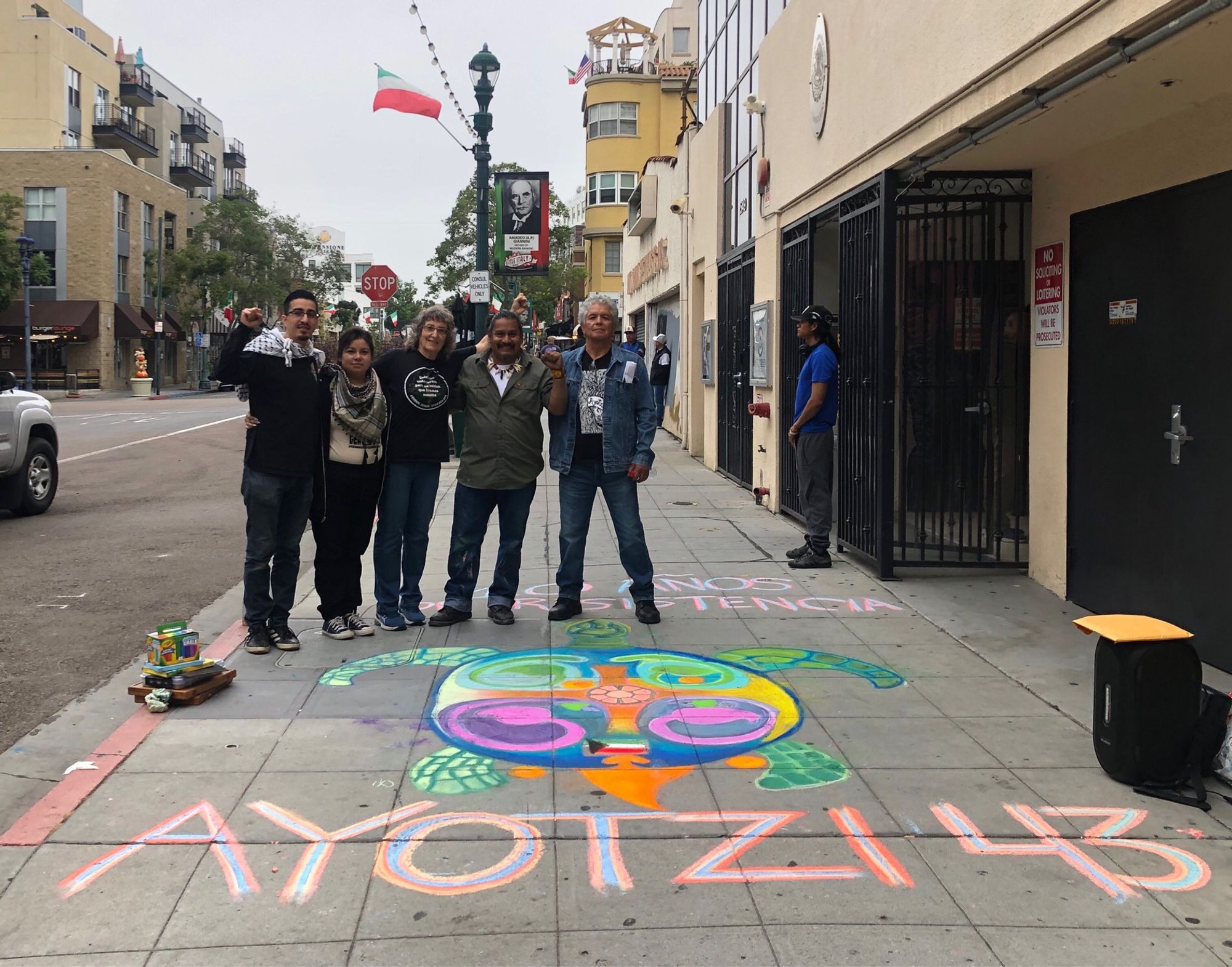 A small group of people standing behind chalk art in front of the Mexican consulate in San Diego. The art commemorates the 43 student teachers from Ayotzinapa who have been missing for ten years as of today.