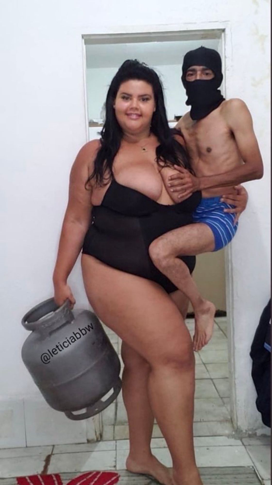 Mulher grande e gostosa de lingerie carregando num braço um botijao de gás e no outro, como um neném de colo, um moço pequeno com balaclava e cueca