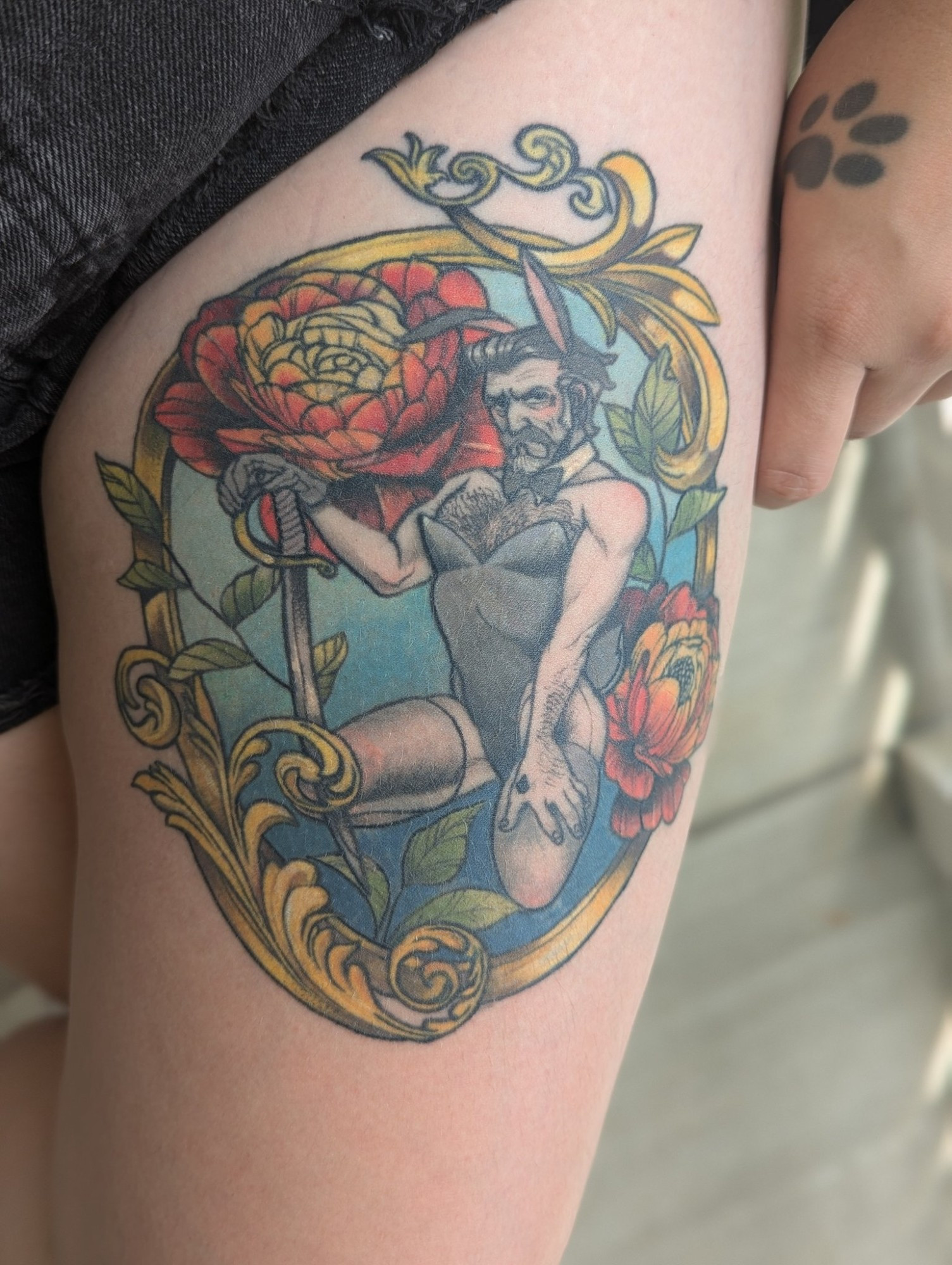 picture of the upper thigh of someone's leg. there is a tattoo on it of Izzy Hands from Our Flag Means Death. He is wearing a bunny girl outfit and bunny ears. He is surrounded by a circular golden frame, and flowers. He is holding a sword and has sheer tights on. 

The person also has a pawprint tattoo visible on their hand resting next to their leg.