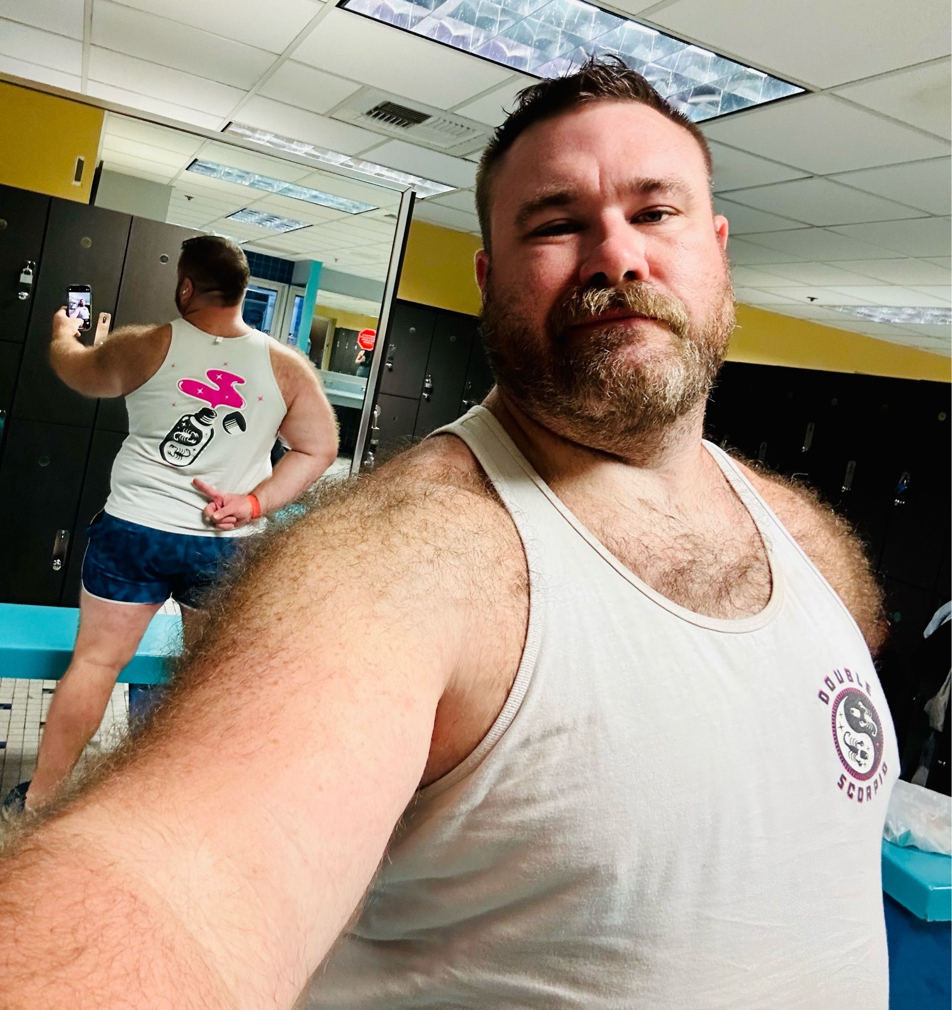 Gaz in a locker room, sporting his white Double Scorpio branded tank top and blue short shorts, facing the camera while using a mirror to point to the large DS open popper bottle logo on his back.