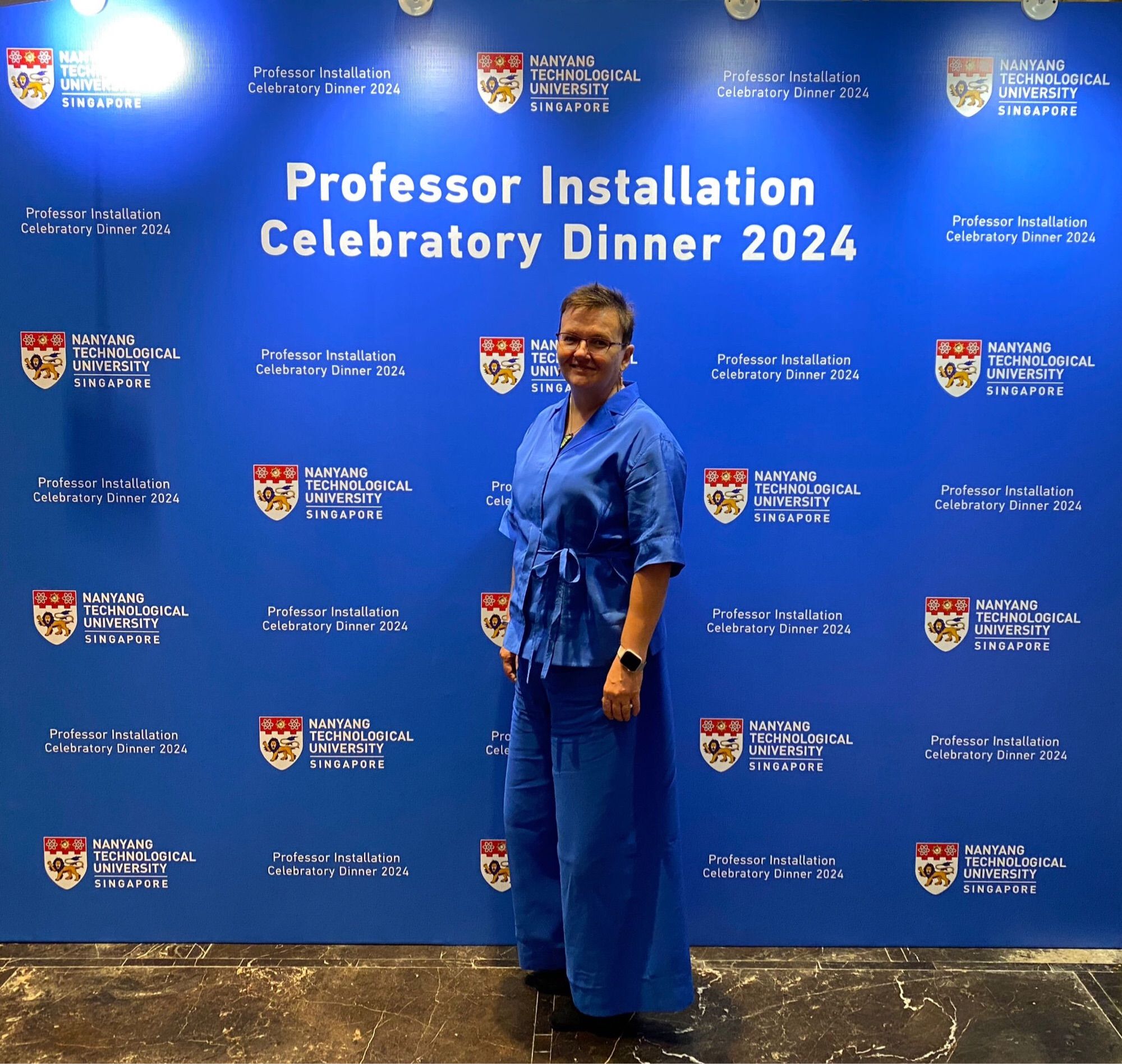 Photo of person dressed in blue pant suit in front of blue wall with university emblems and signage