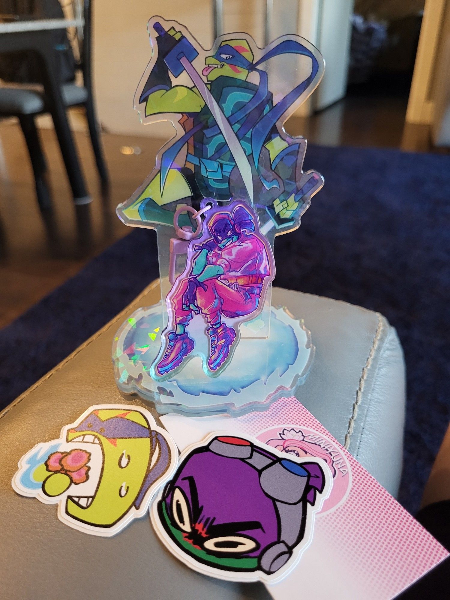 the previous mentioned standee and keychain shown together along with the creator's card which reads, "Miniyuna." there are also two freebie stickers, one of Dontello's face looking very angry in a cartoonish style and another with Leonardo eating pizza puffs in a cartoonish style.