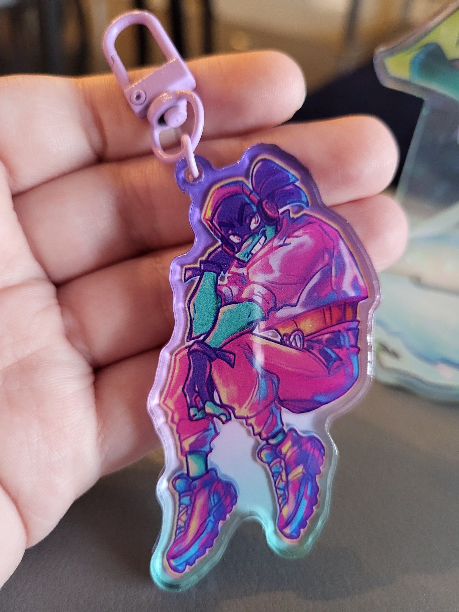 a keychain of Donatello from Rise of the TMNT. he is sitting down with one hand propped up by his head while the other rests over his knee. he is wearing a lighter pink cropped shirt with slightly puffy sleeves that end half way down his arms. his pants are a darker pink color while his tennis shoes and gloves are purple like his signature mask.