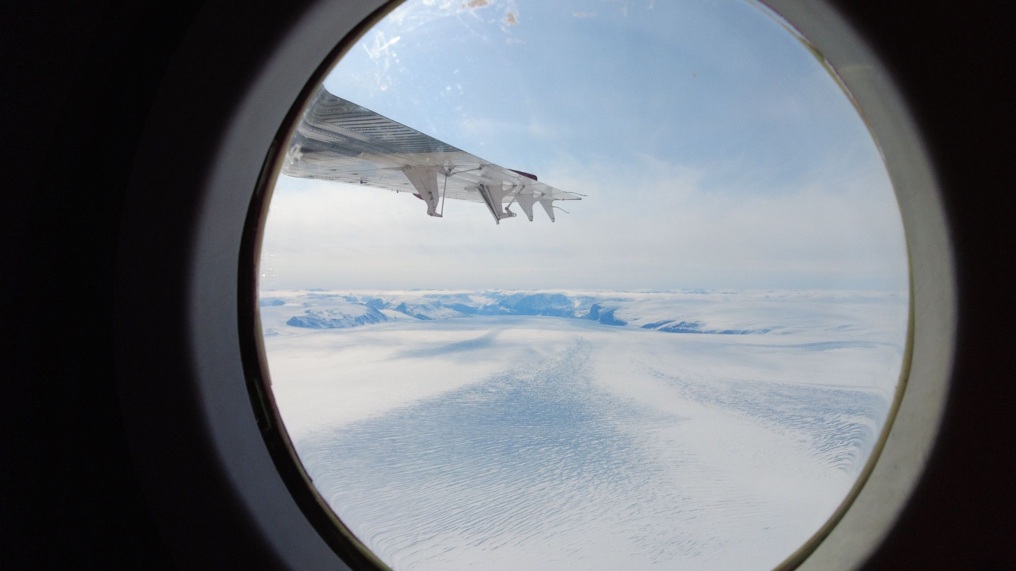 View from a circular window with lightly clouded blue skies, an airplane wing, and white, crevassed glacier surface and mountains in the background.