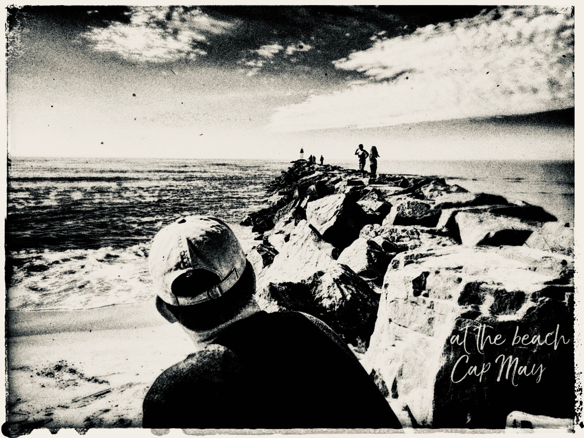 schwarzweiß Bild zeigt Mann wie er an einer Mole sitzt. Im Hintergrund sind zwei Kinder zu sehen die auf dieser aus großen Quader gebauten Mole stehen. Das Bild heißt At the beach - Cap May