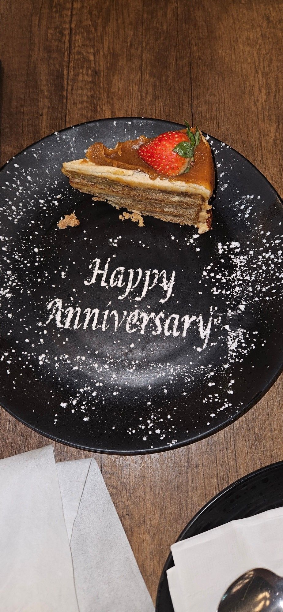 Black plate on wooden table dusted with Icing Sugar (powdered sugar) features a design that says "Happy Anniversary".

A layered cake sits on the upper middle of the plate with a strawberry placed on top.

Apple Mango Habernero Cake