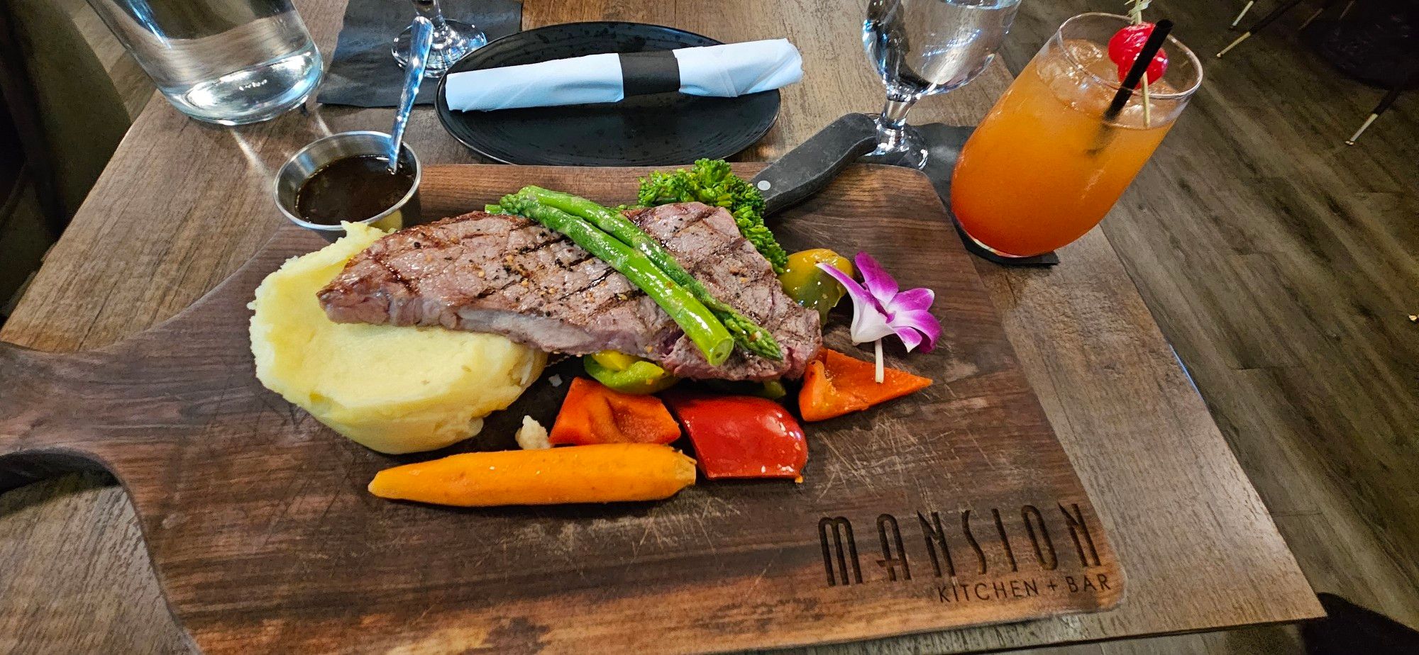 Wooden chopping board on wooden table.

Wooden chopping board used as a "plate" with mashed potatoes, steak, flowering broccoli, asparagus, a carrot, pieces of mixed bell peppers.

A single purple and white orchid blossom is on the middle right of the picture on the board.

Upper right hand corner of the picture features an acoholic drink (Mai Tai) with red and orange colours and a cocktail stick placed over the top of the glass with 2 Maraschino Cherries.