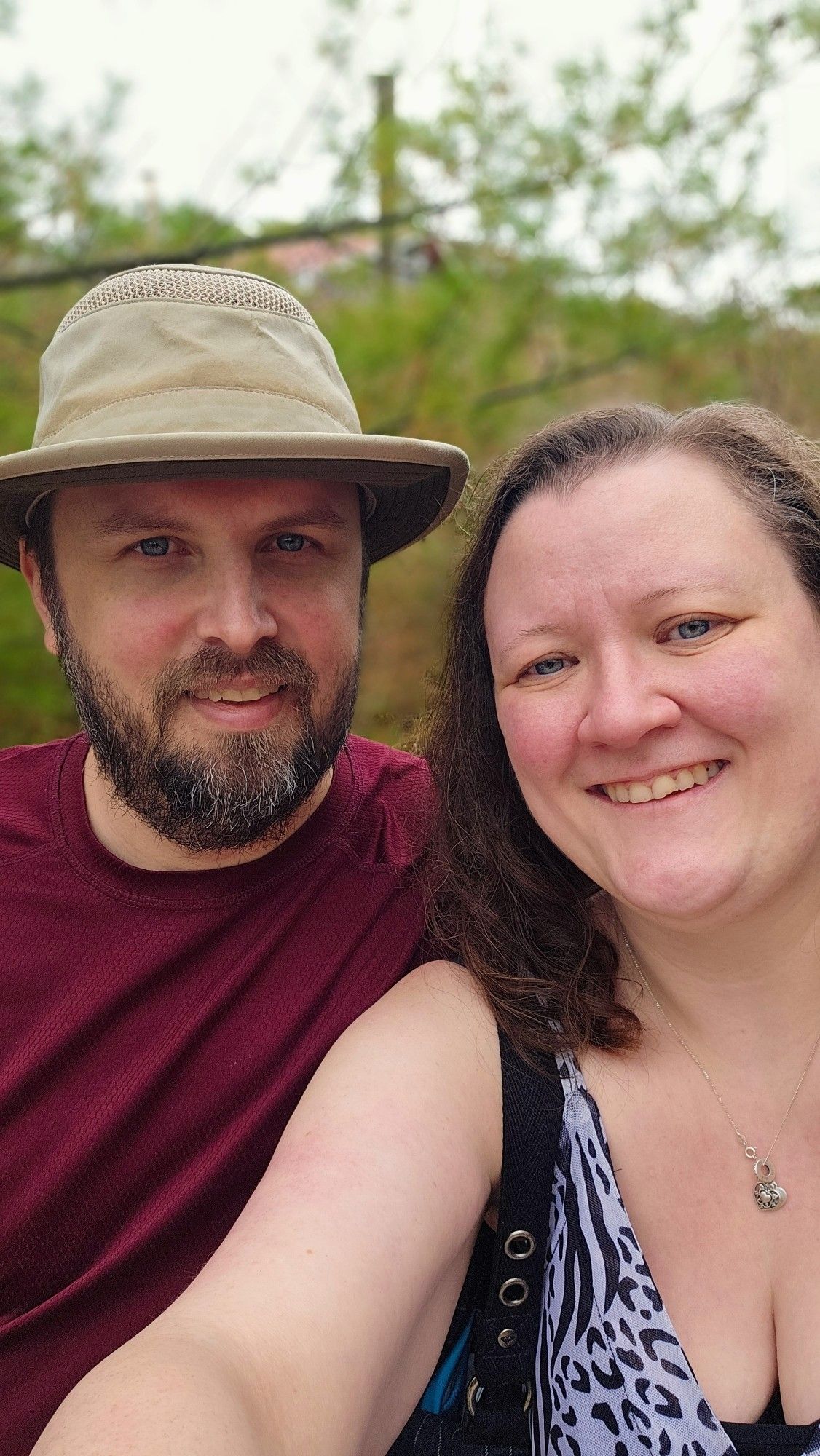 Picture of a man (left in picture) in a red shirt and light khaki hat with a slight smile on his face and a woman in a snow leopard print dress with a smile (right in pic) with a blurred background of leaves and other foliage.