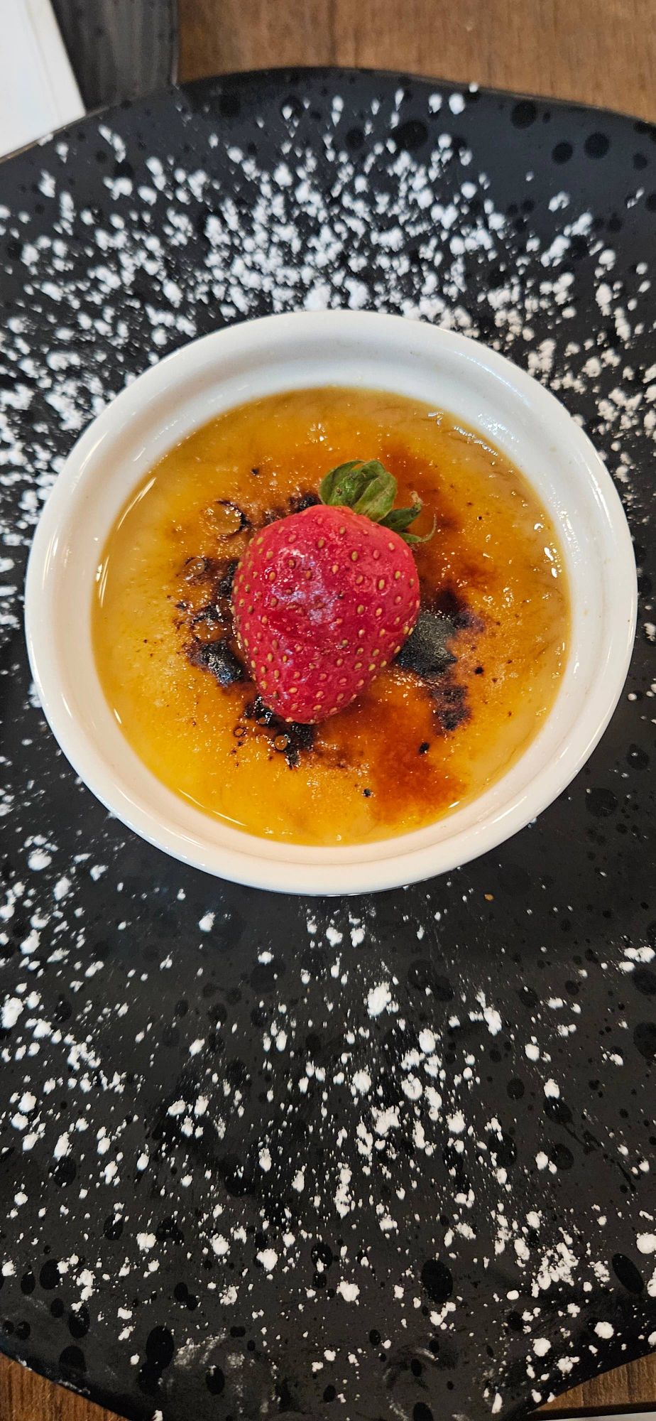 A black plate dusted with icing sugar (sugar powder) on a wooden table. 
A white ramekin sits in the middle of the plate with a strawberry on top of the melted sugar topping of a Crème Brûlée.