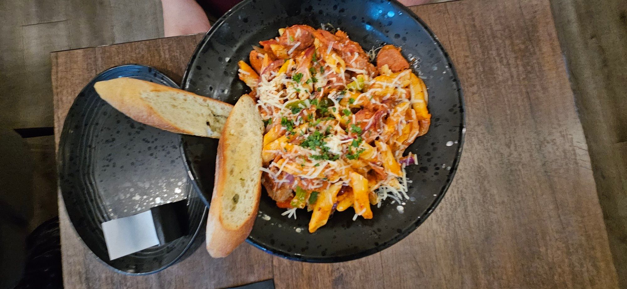 Black bowl on wooden table. 

2 pieces of Garlic Bread rest on the left side of the bowl in picture. 

Black bowl is full of Arrabbiata Spiced penne pasta with Spicy Italian Sausage.