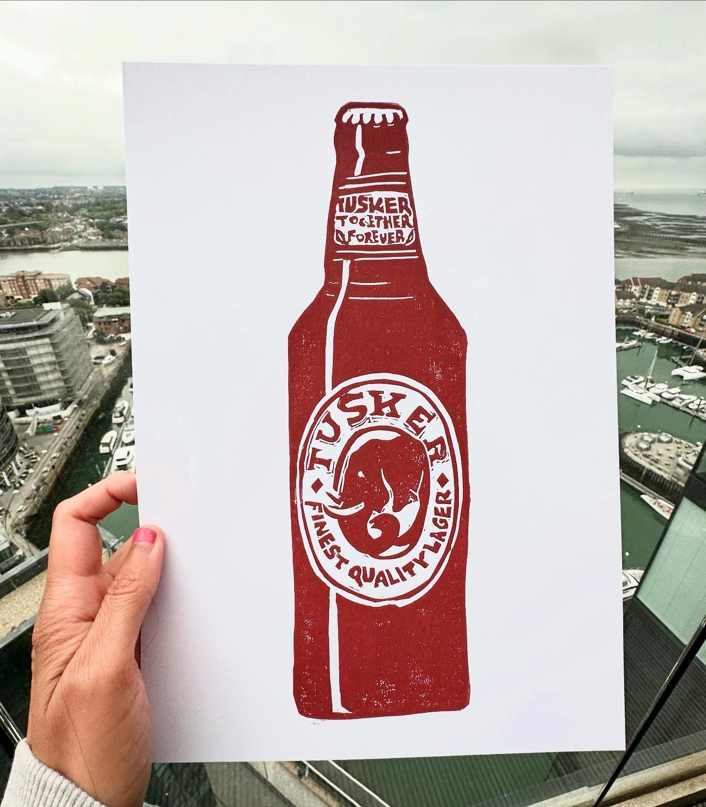 A hand holding an art work Lino print of Tusker Beer bottle in burnt Sienna. The background is a marina setting in Southampton.