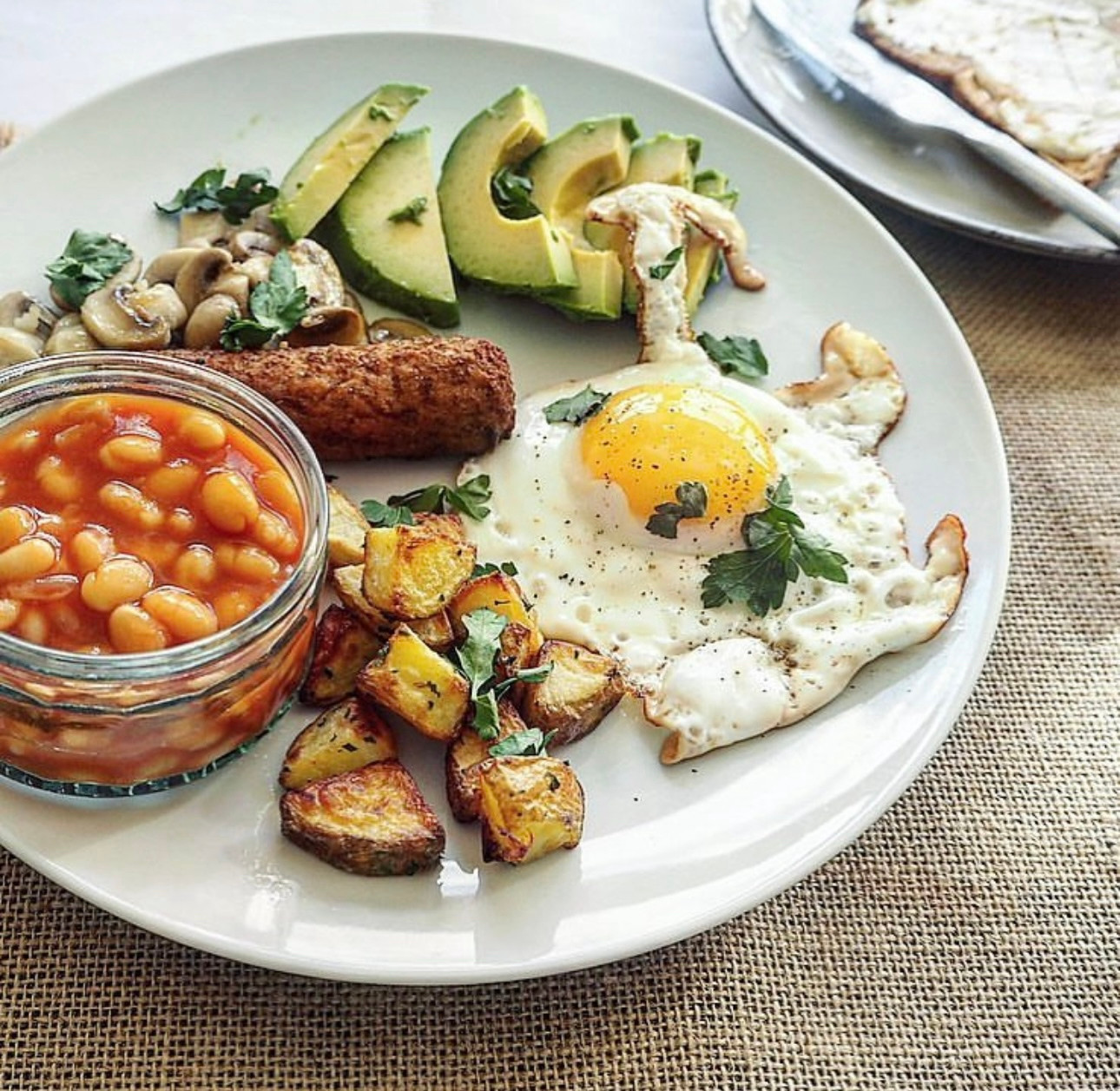 A veggie breakfast including a fried egg, mini roasted potatoes, a vegetarian sausage, sliced avocado, garlic baby mushrooms, and a ramekin ok baked beans. There’s a plate of toast and a butter knife slightly out of shot too.