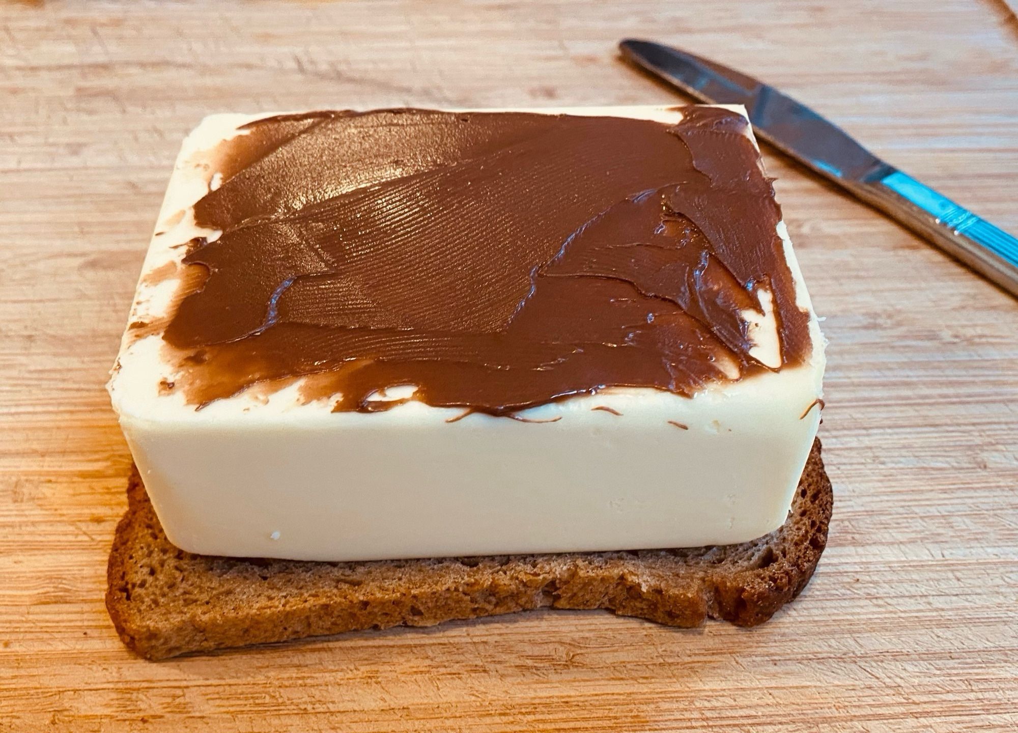 Eine Scheibe Brot, darauf ein großes Stück Butter (250g), darauf ein Hauch Haselnusscreme. Im Hintergrund liegt ein Messer, alles zusammen liegt auf einem Holzbrett