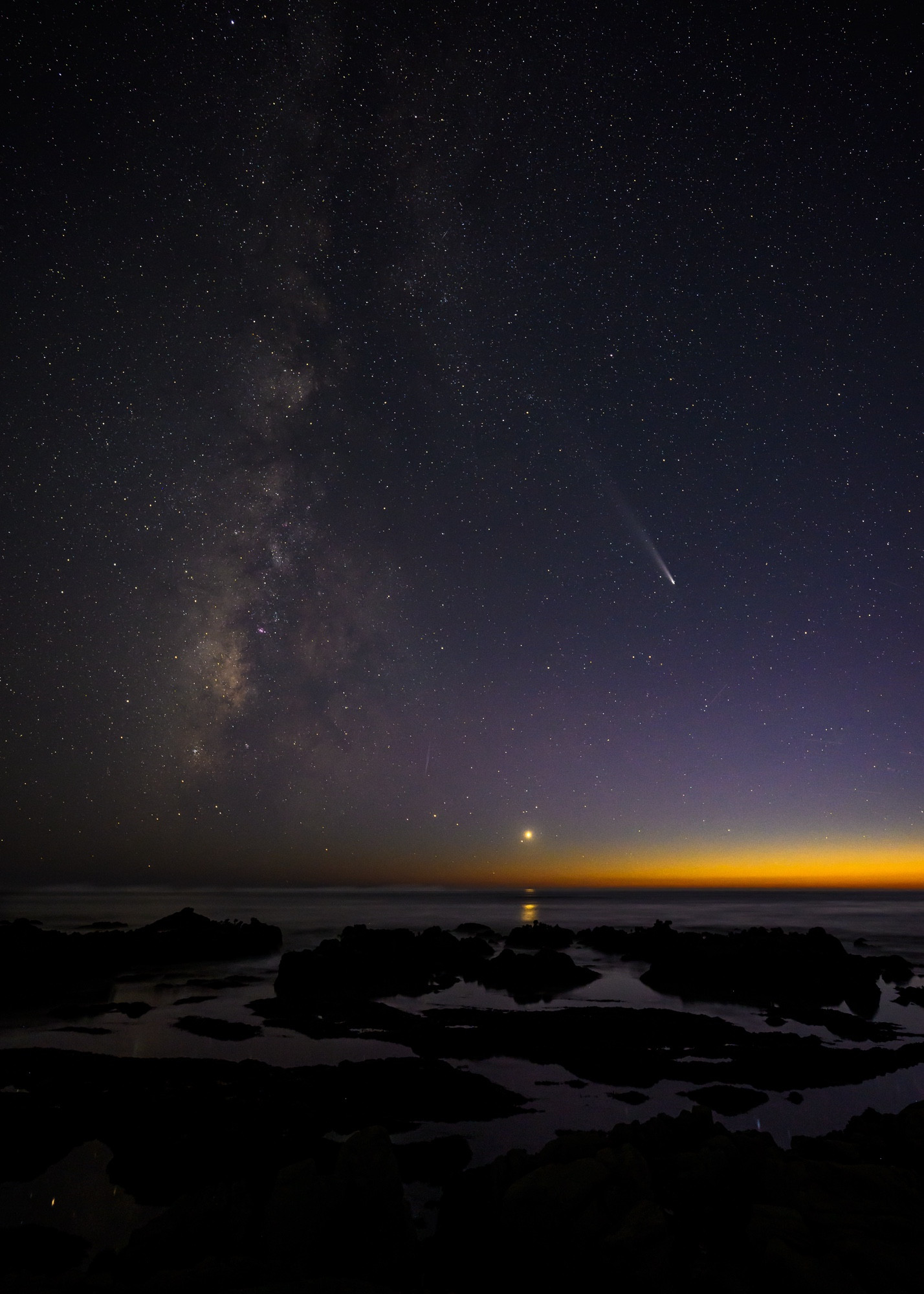 A star-filled sky over the coast. On the right, a comet pointed down. On the left the Milky Way. In between, Venus whose reflection can be seen in the water. The remnants of sunset are on the horizon.