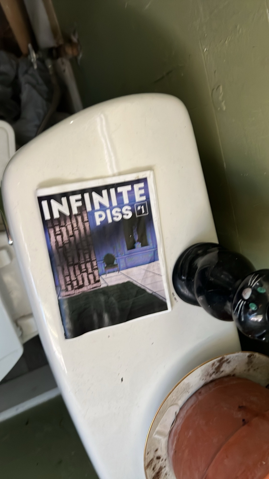 a zine sits on a toilet, reading “infinite piss” on the cover, maybe an allusion to infinite jest?