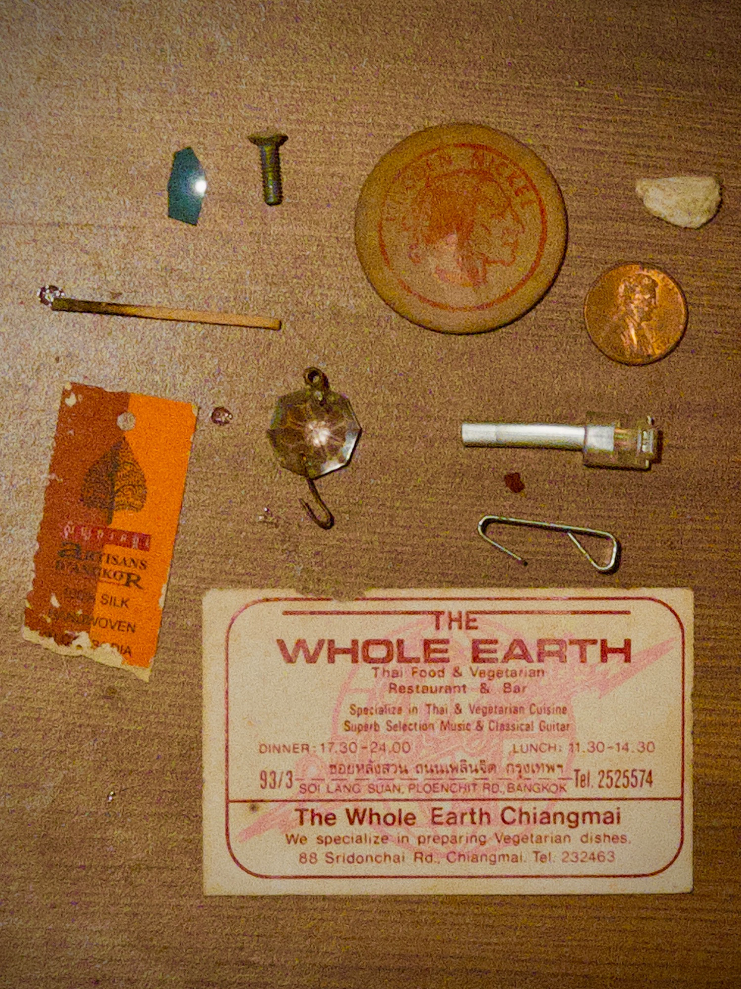 contents of an old wooden side table. a tag for a silk scarf, the end of a telephone wire, a penny, a ball of washed paper, a screw, a shard of translucent blue plastic, a hook, a burnt match, a plastic jewel with a hook in it, and a business card for The Whole Earth Thai Food and Vegetarian Restaurant and Bar, Soi Lang Suan, Ploenchit Rd., Bangkok