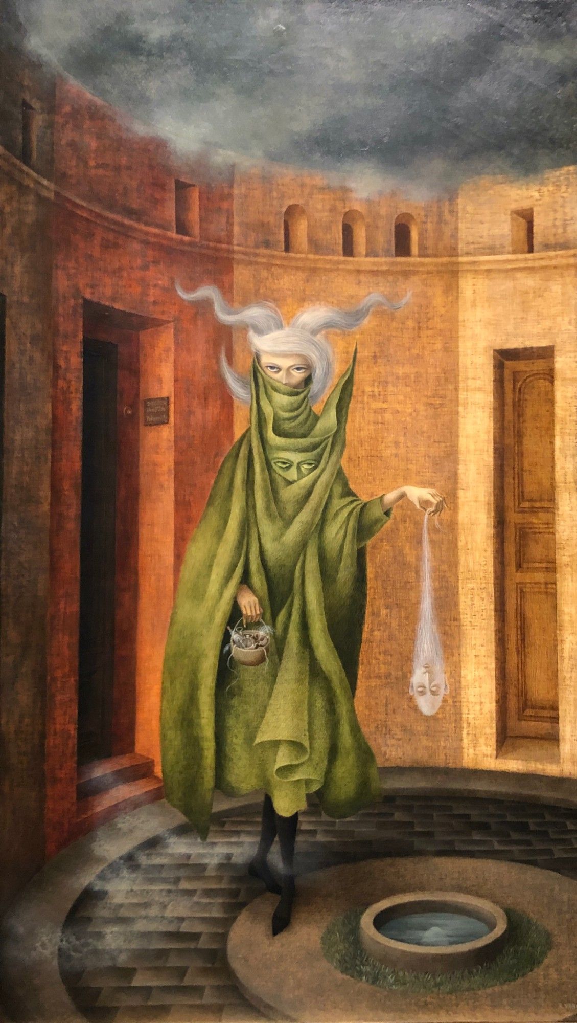 A woman with white hair that floats out in two horn-like protrusions and wearing a green cloak with a face in it exits a door holding a man's head in her left hand and a basket in her right hand. The man's head has a long beard, and the woman holds him disdainfully by the tip of the beard. She appears to be about to drop him into a circular pool of water in the courtyard she's in.