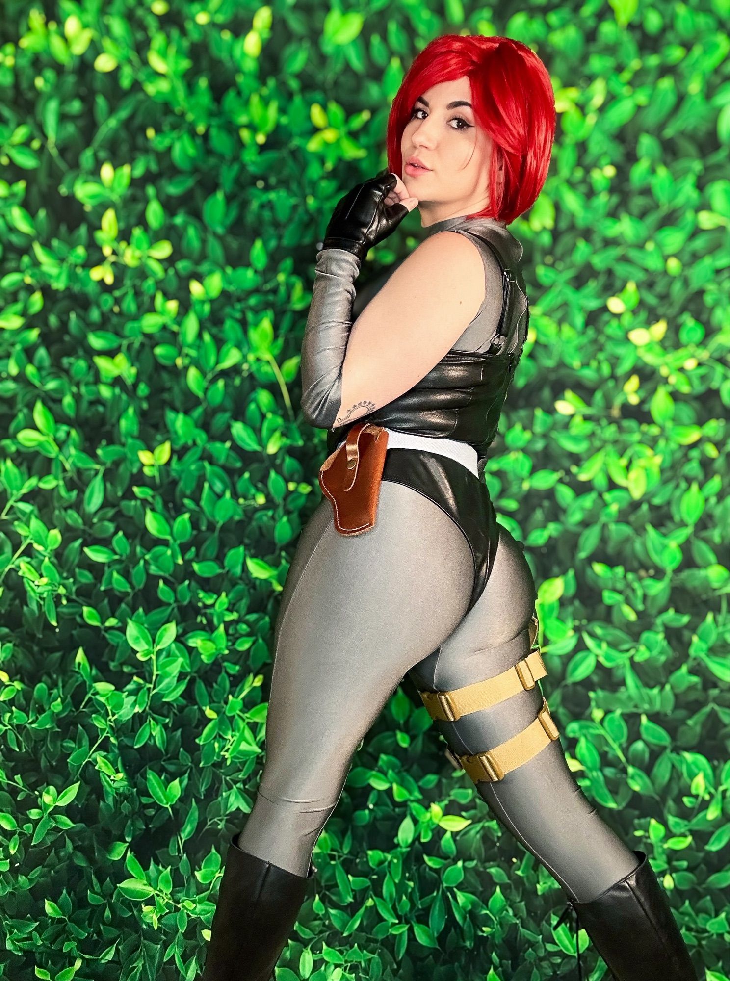 photo of bloodyfaster in cosplay as regina from dino crisis in front of a green leafy background. her back is turned and she is looking over her shoulder into the camera