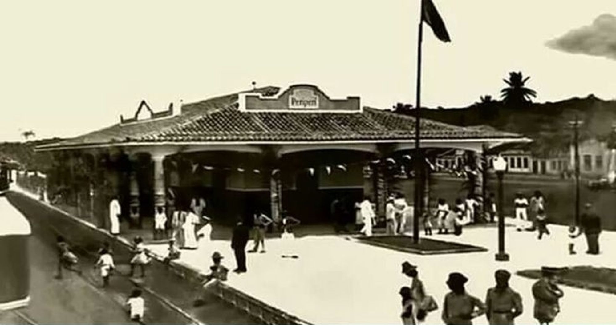 Foto, em preto e branco, da estação Periperi onde aparece em destaque a plataforma de embarque, ao centro, com a  cobertura de telhado e o nome da estação no alto do mesmo. E na plataforma passageiros aguardando o trem. Ao fundo da estação algumas casas do bairro.