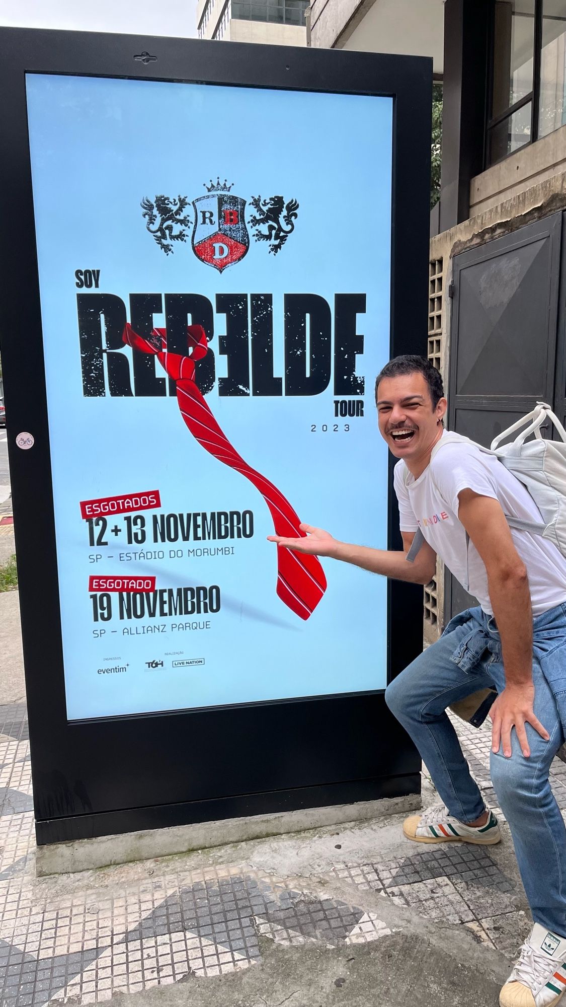 Matheus Mesquita (@mesquitavs) junto a letreiro de rua anunciando os shows da Soy Rebelde Tour em São Paulo.