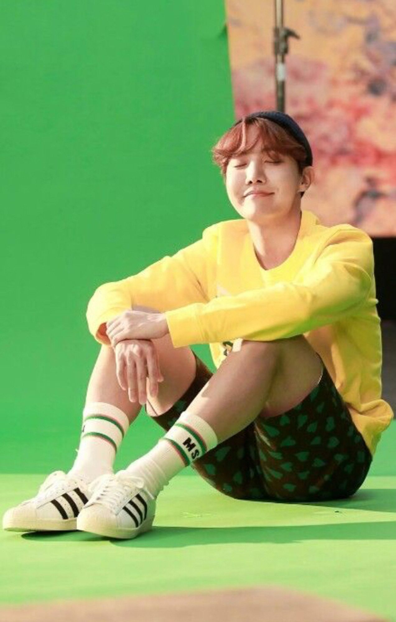 j-hope is sitting on the floor and resting his arms to his knees. He’s smiling with his eyes closed and it looks like he’s basking in the sun even though he’s in a studio with a green background. He’s wearing shorts and sport socks and sneakers and her legs are very pretty. His top is a yellow sweatshirt and his wavy hair is auburn.