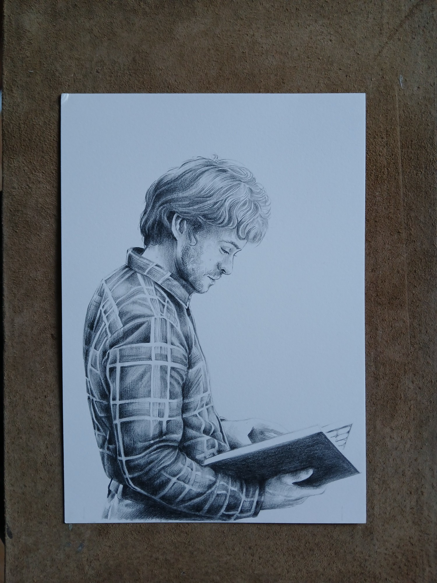 Pencil drawing on paper of a curly haired Will Graham in a plaid shirt. He is looking down at the book he is holding in his hands.