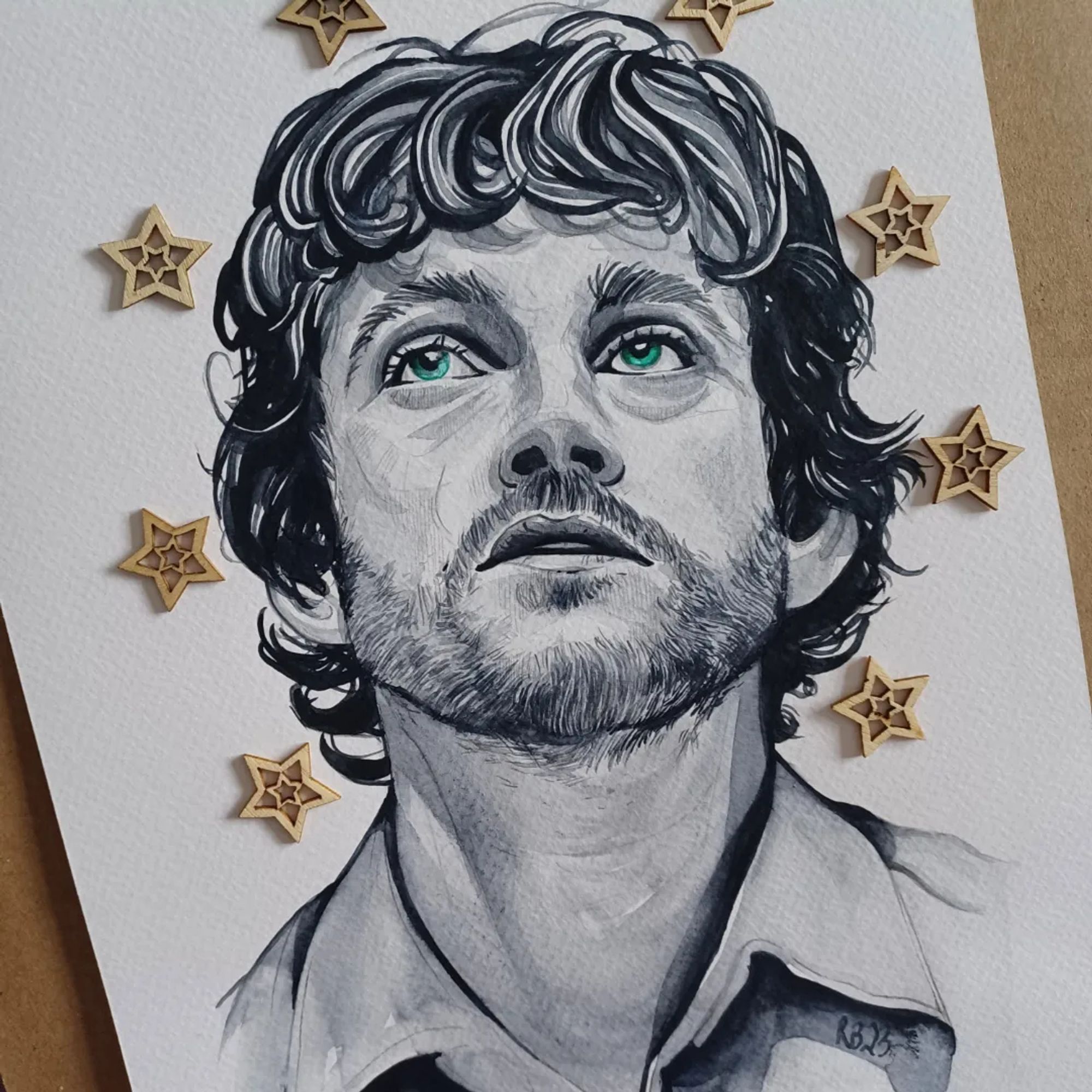 Watercolour drawing of Will Graham on paper. His eyes are brilliant green. There are wooden stars on the page placed around his head.