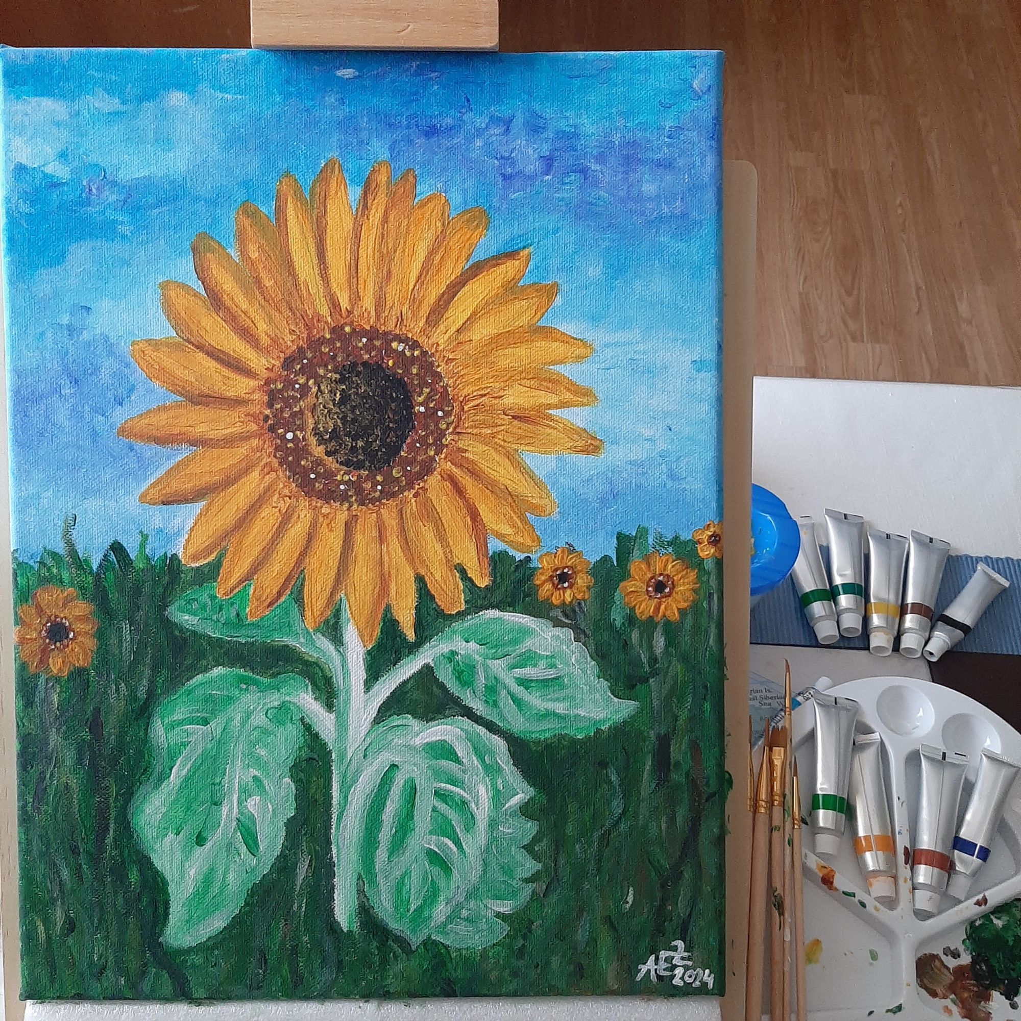 Foto von einem Acrylbild auf einer Staffelei, das eine große Sonnenblume zeigt. Daneben rechts auf dem Tisch liegen mehrere Farbtuben, Pinsel und andere Malutensilien.