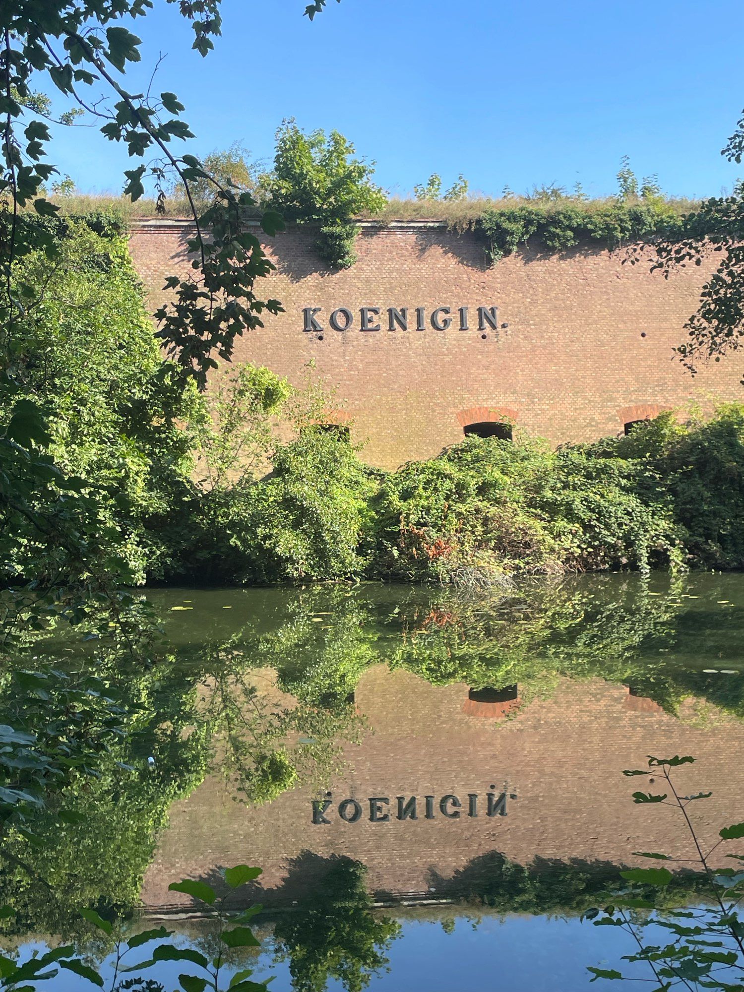 Die Bewehrung der Spandauer Zitadelle mit Schriftzug „Königin“, davor der Burggraben, in dem sich der Schriftzug und die Begrünung spiegeln