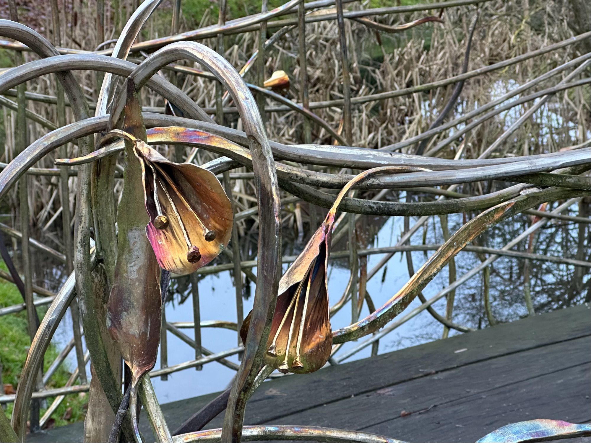 Von einem Künstler geschmiedetes Gitter, das als Begrenzung einer kleinen Holzbrücke dient. Das Gitter ist in Wellen gearbeitet und hat auch große Blüten, die golden schimmern.