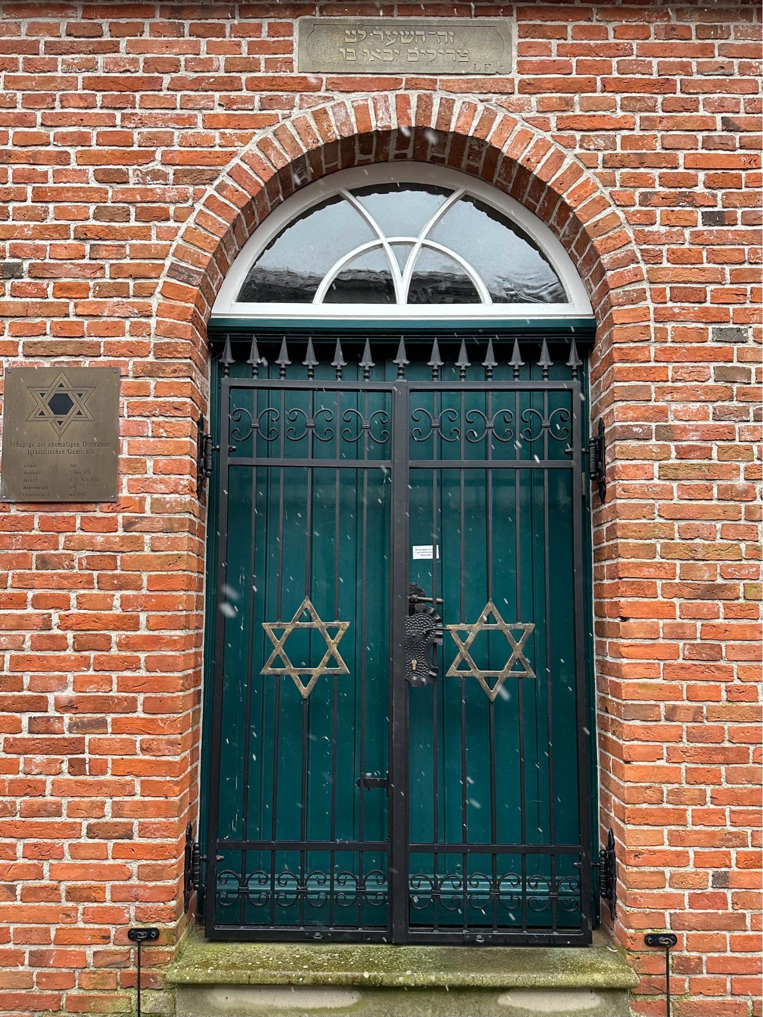 Eine grüne Eingangstür mit halbrundem Oberlicht, die in ein Backsteingebäude führt. Die Tür ist mit einem schwarzen Gitter geschützt, in das zwei goldene Davidsterne eingearbeitet sind. Es handelt sich um den Eingang der ehemaligen Synagoge in Dornum.