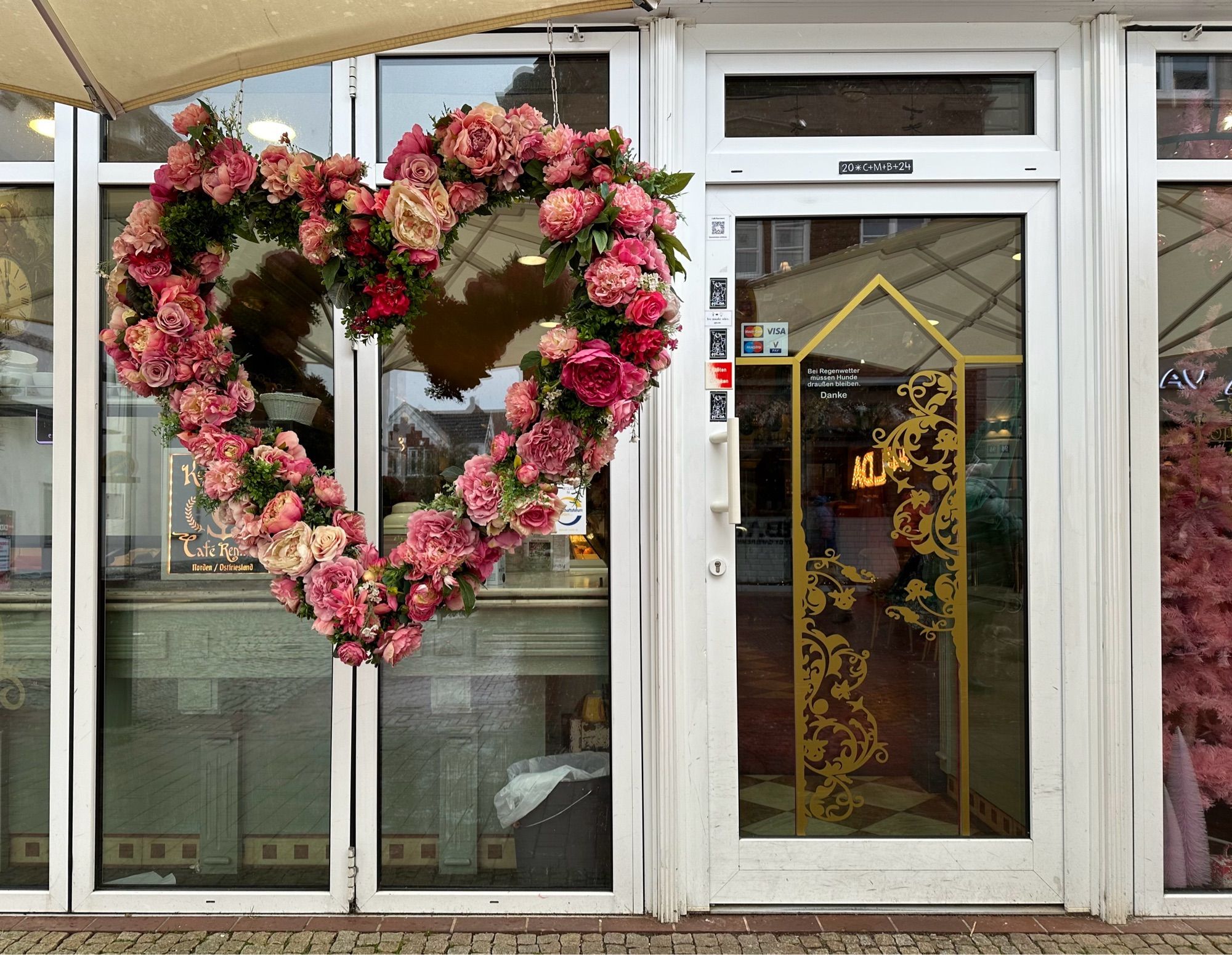 Eine gewöhnliche Glastür, die in ein Cafe führt, aber vor dem Schaufenster links davon hängt ein großes Herz aus Rosen