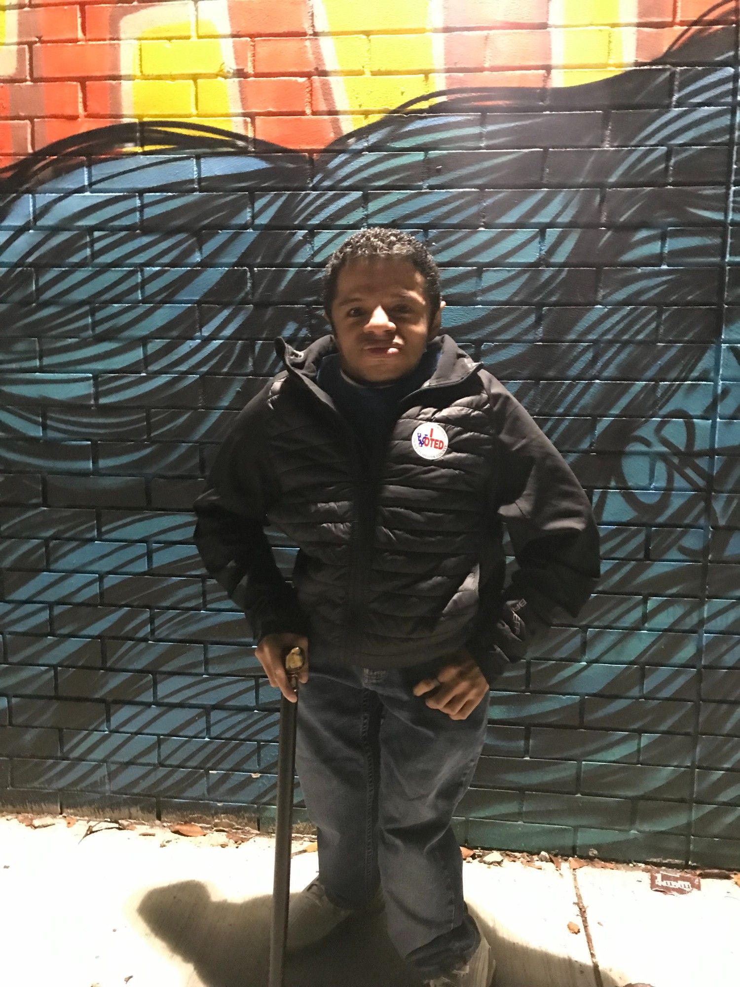 A pic of me, a Little Person disabled Latino male, standing in front of a mural at my local polling location on election day. A sticker that says "I Voted" on my black bubble jacket.