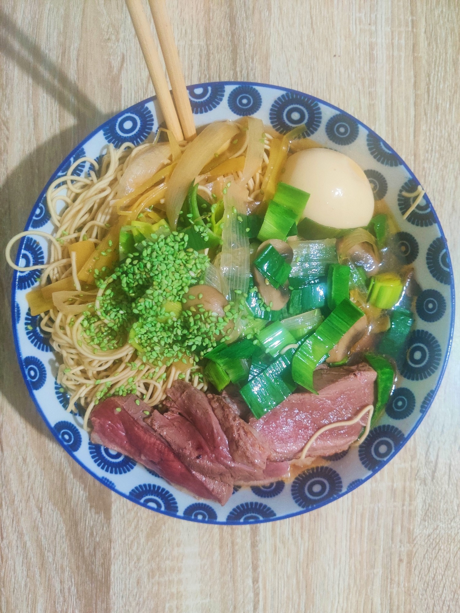 Ramens dit "canarticho" nouilles, sésame au wasabi, poireau vert, viande de canard bien rouge, oeuf mollet mariné au soja, huile du bouillon bien brillante mais à peine visible tant y a de trucs dans le bol bleu. Une paire de baguettes est trempée dans le bol, tout est prêt pour déguster.