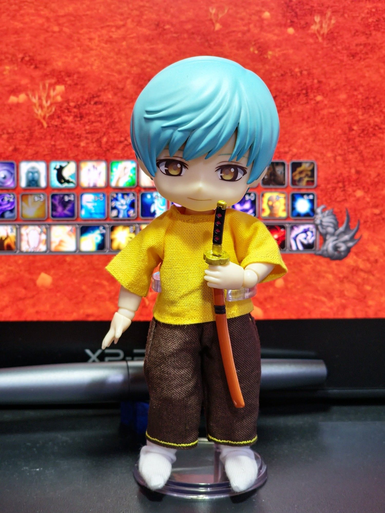 A 1 12th scale ball jointed doll with short blue hair. They wear a bright yellow t-shirt and brown trousers with yellow thread detail. In their left hand, they hold a sheathed katana close to their chest.