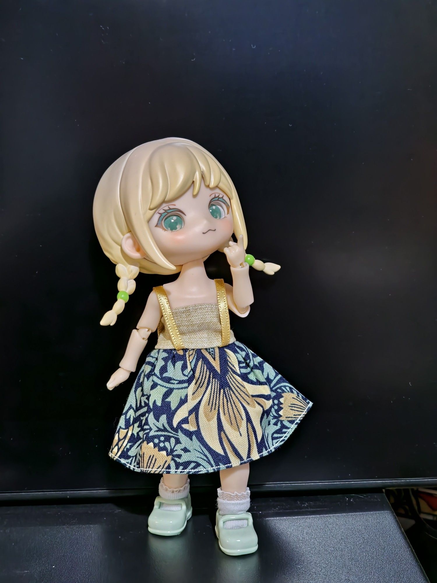 A blonde ball jointed doll posed standing with a thoughtful expression, head titled upwards. She is wearing a dress with a plain beige bodice with gold ribbon straps and William Morris skirt of bold floral design in beige, mint green and navy blue