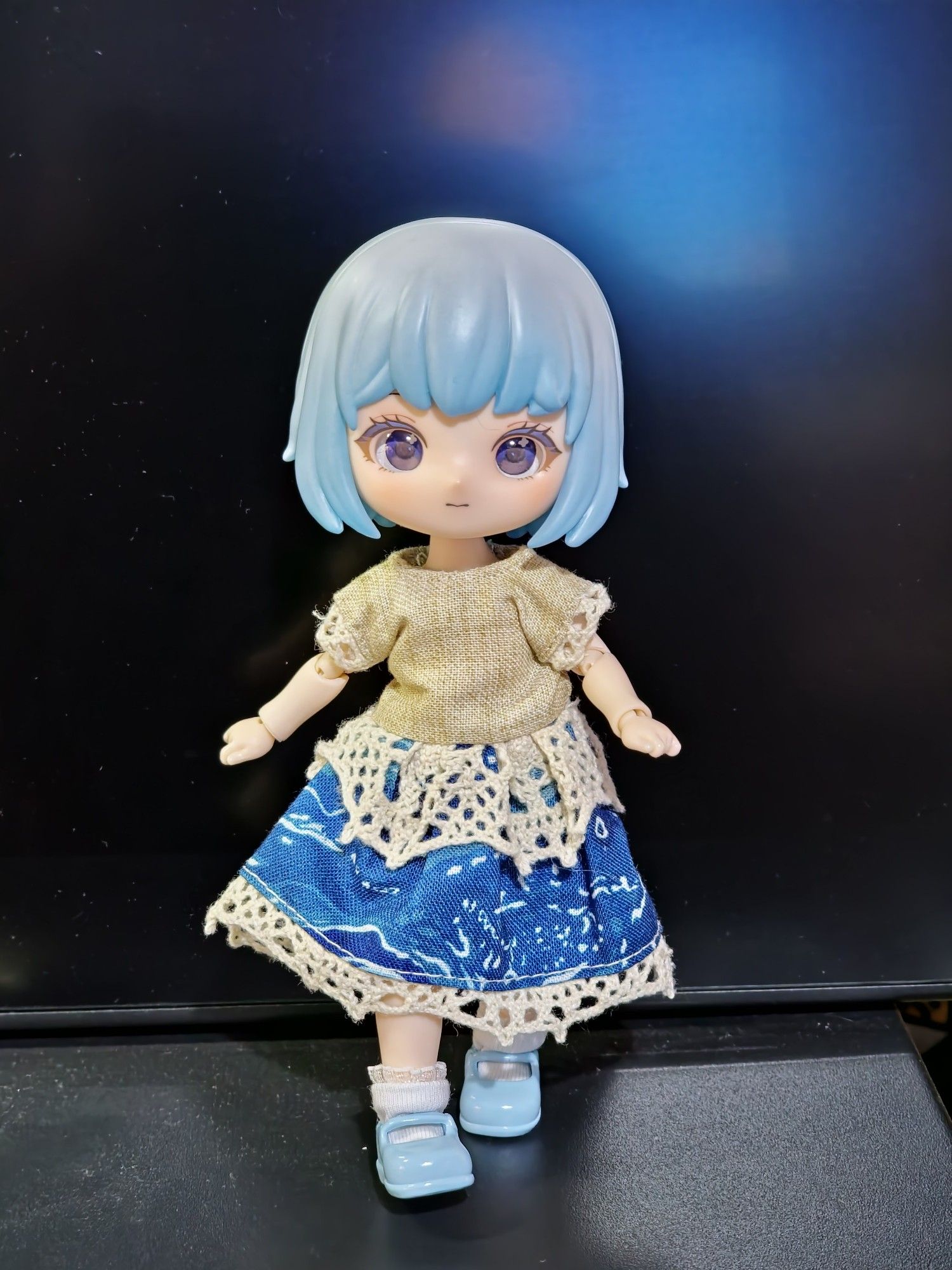 A blue-haired ball jointed doll walking towards the camera like fashion model. She is wearing a mid length dress with beige, bodice with lace on the short sleeves. The blue skirt of the dress has a water swirl design and is fringed with and overlayed with lace that looks like sea foam