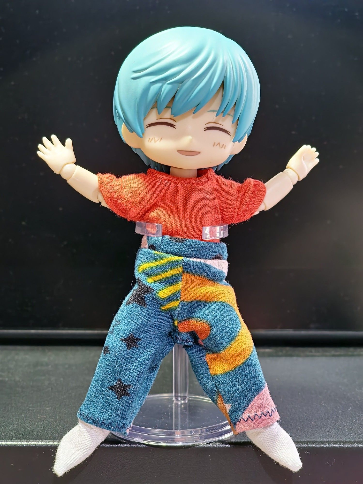 A 1 12th scale ball jointed doll with short blue hair is doing a star jump. Their eyes are shut whilst smiling and blushing. They are wearing a short sleeved red top and pyjama bottoms that are dark turquoise decorated with orange and pink swirls and black stars.