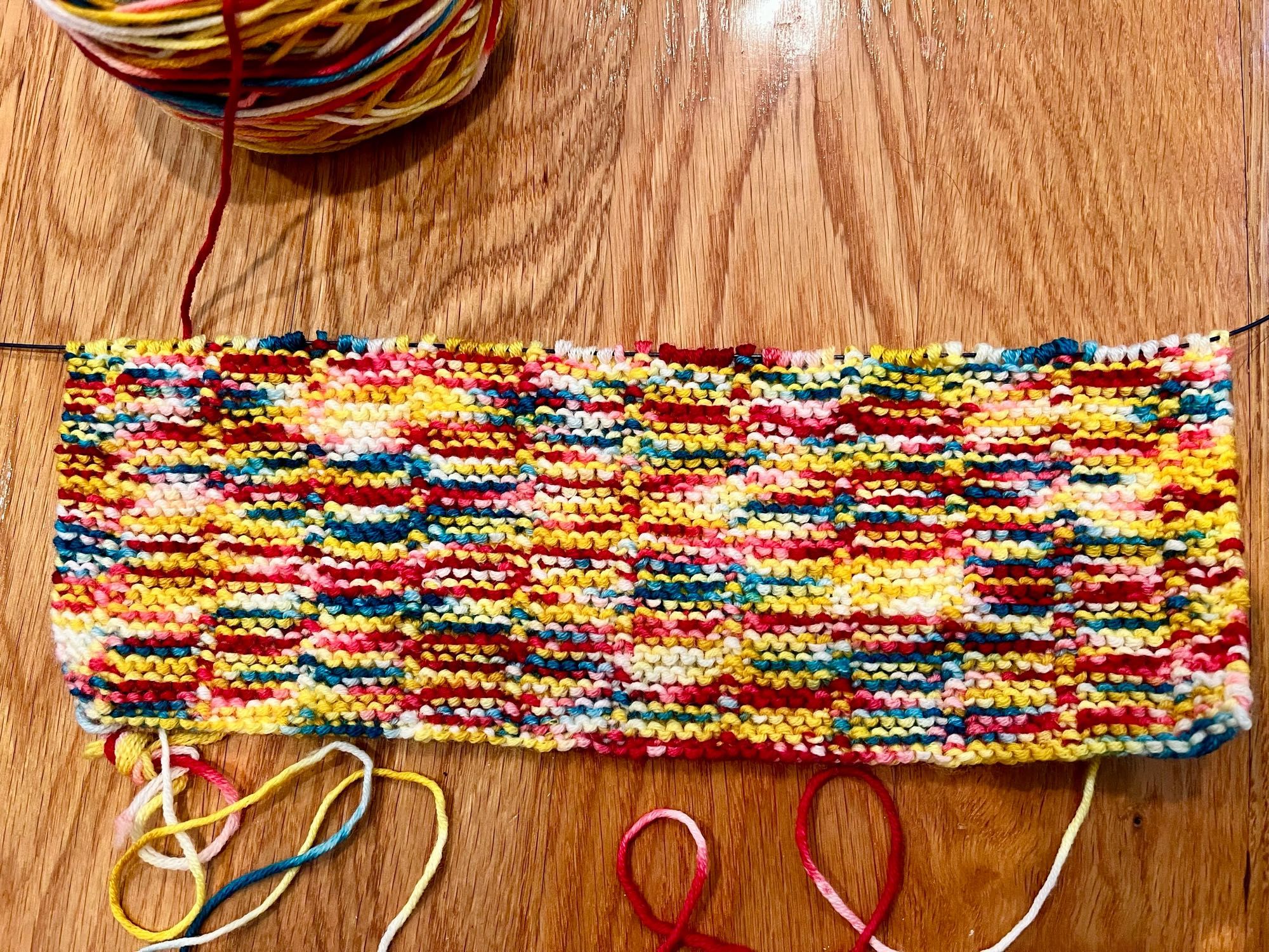 About 30-40 rows of a merino wool scarf in the Agolant colorway from the Republic of Yarnia, pictured on a wood floor. The color variegation here working much better imo than the last attempt at this project.