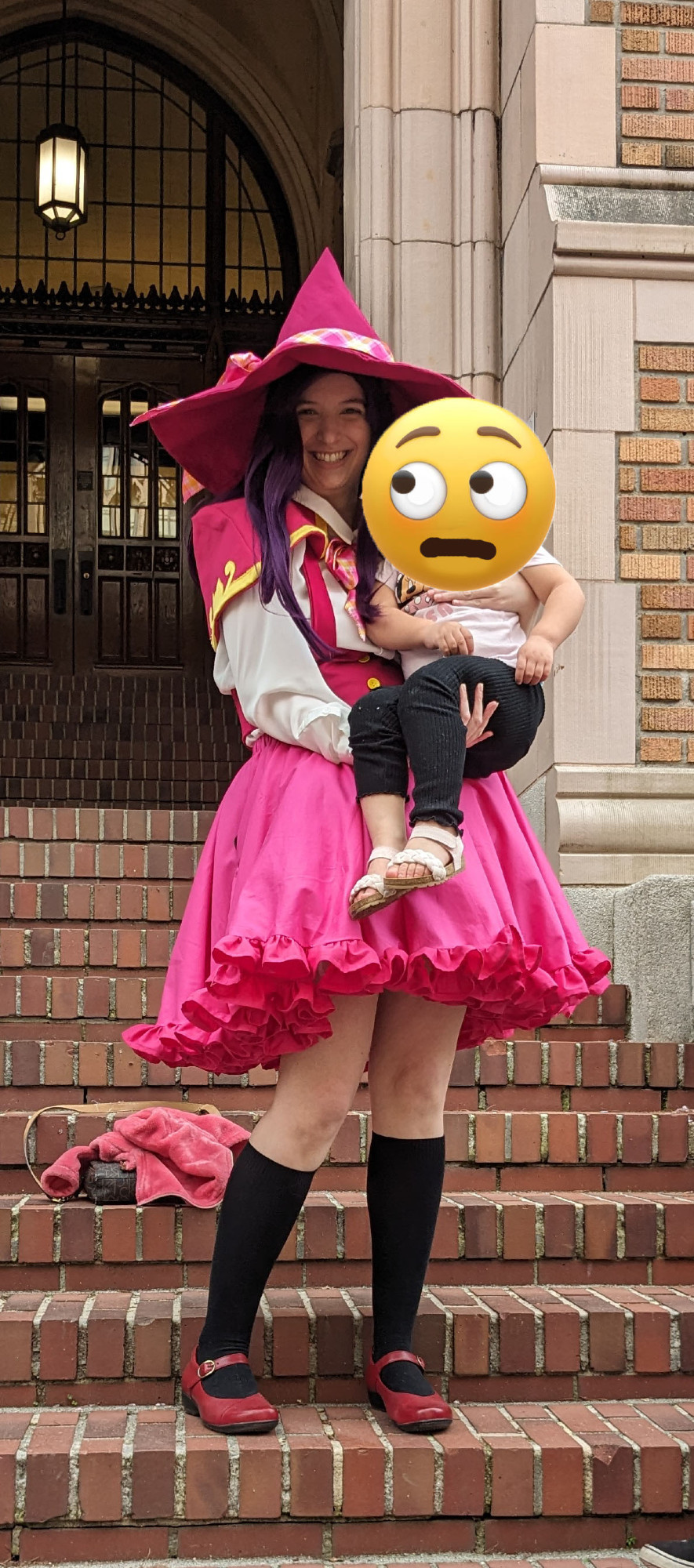 A cosplayer dressed as Liko Izayoi awkwardly holding a child. The child's face has been replaced with an emoji meant to convey "I'm not sure about this person."