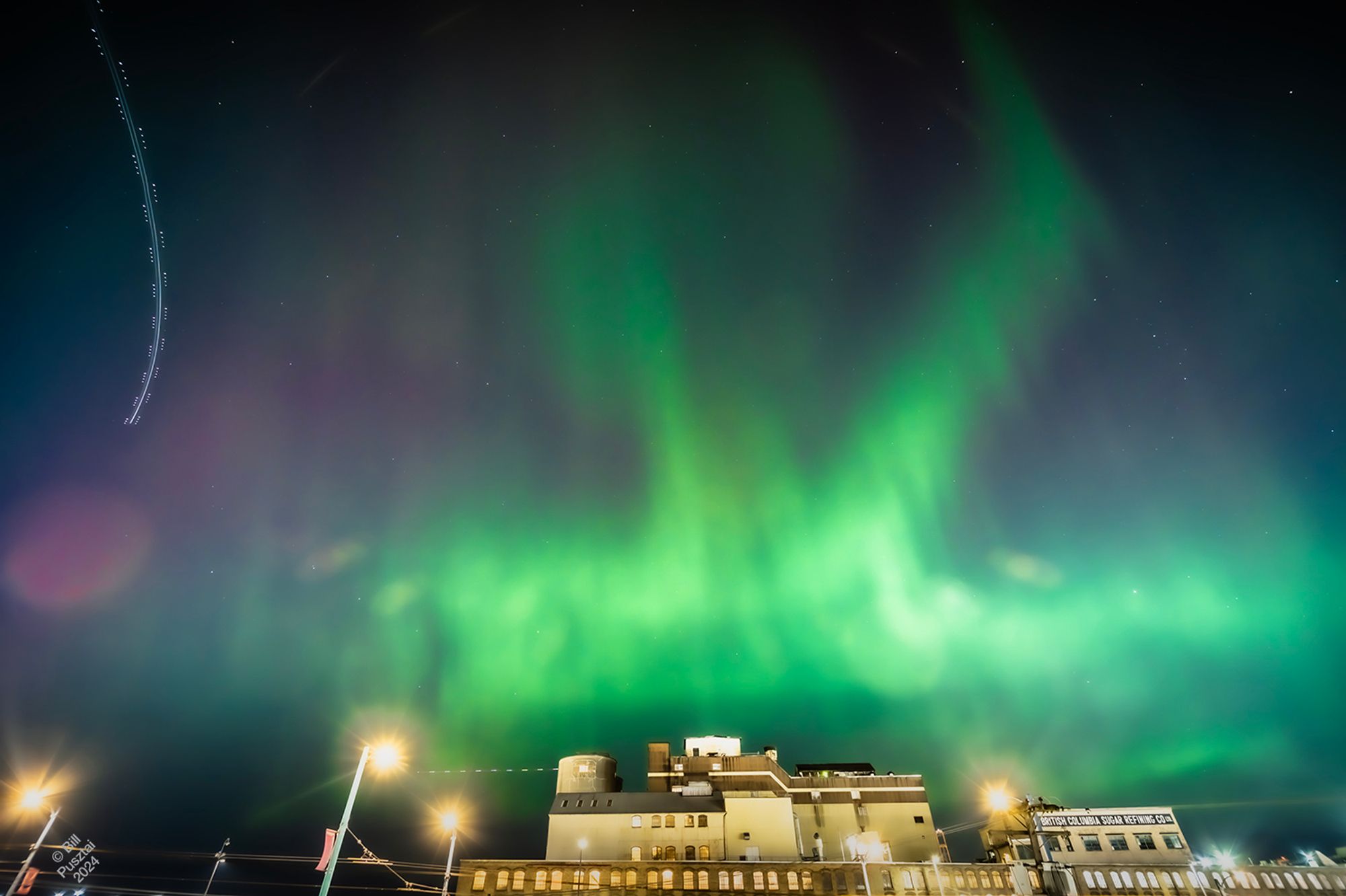 Colour photo, 13 second exposure. In the bottom 1/4 a brightly lit industrial building (the refinery). In the top 3/4 the sky with aurora, mostly bright green with patches of magenta. Top left and bottom center, the light trails of planes. Throughout, lens flare light artefacts. Top 2/3, stars through the lights.