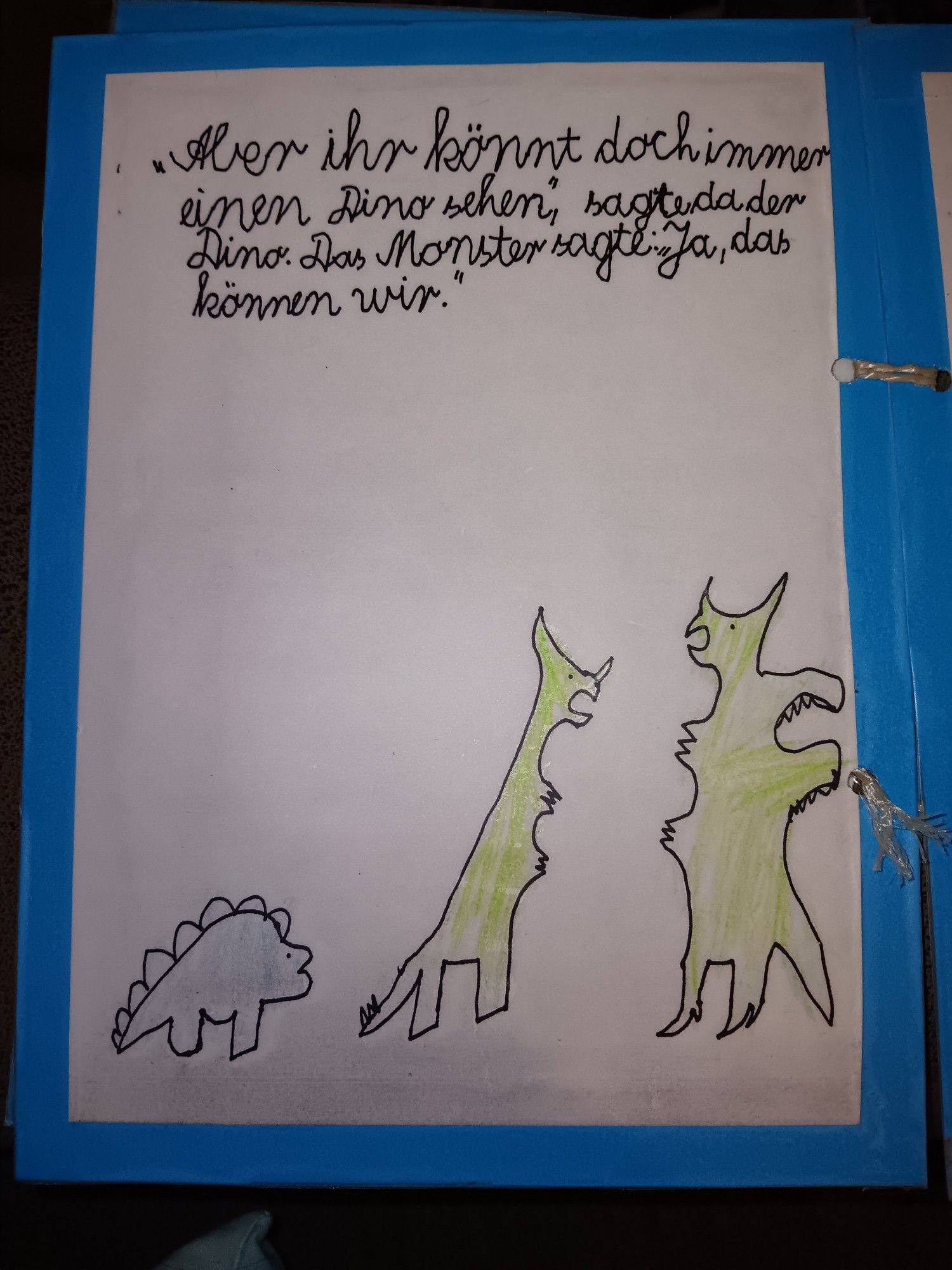 Seite eines selbst gebastelten Kinderbuchs mit einer murkeligen Kinderzeichnung auf Tonpapier hinter Folie. Der Dino und das Monster sehen den Drachen an. Text: "Aber ihr könnt doch immer einen Dino sehen, " sagte da der Dino. Das Monster sagte: "Ja, das können wir."