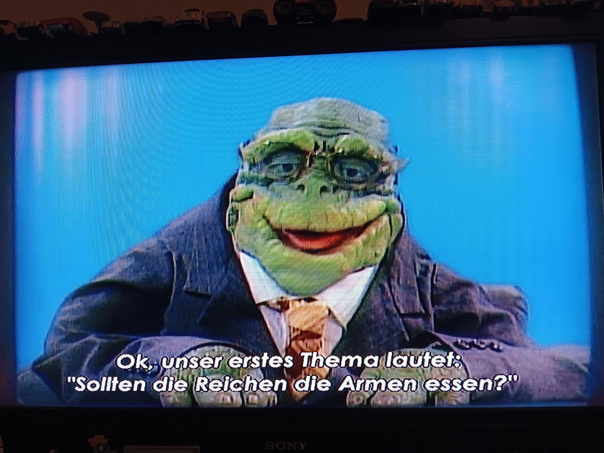 Szene aus der Puppensitcom DIE DINOS: ein Talkshowmoderator, der ein anthropomorpher Dinosauriermann im Anzug ist, sagt: "OK, unser erstes Thema lautet: "Sollten die Reichen die Armen essen?""