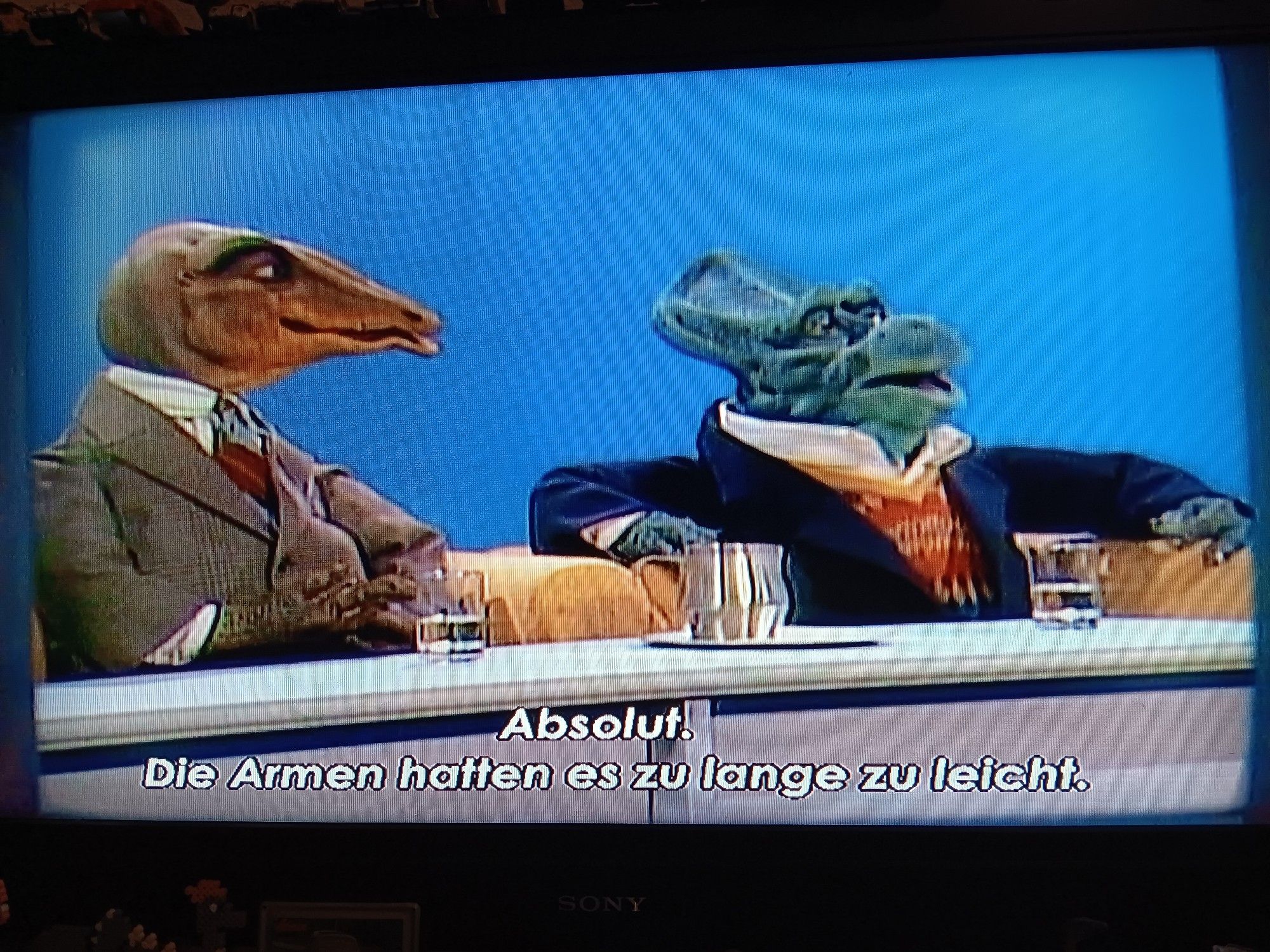 Szene aus der Puppensitcom DIE DINOS: zwei anthropomorphe Dinosauriermänner, ein Gallimimus und ein Protoceratops, die beide in scheinbar teure Klamotten gehüllt sind, sitzen nebeneinander. Der Protoceratopsmann sagt: "Absolut. Die Armen hatten es zu lange zu leicht."