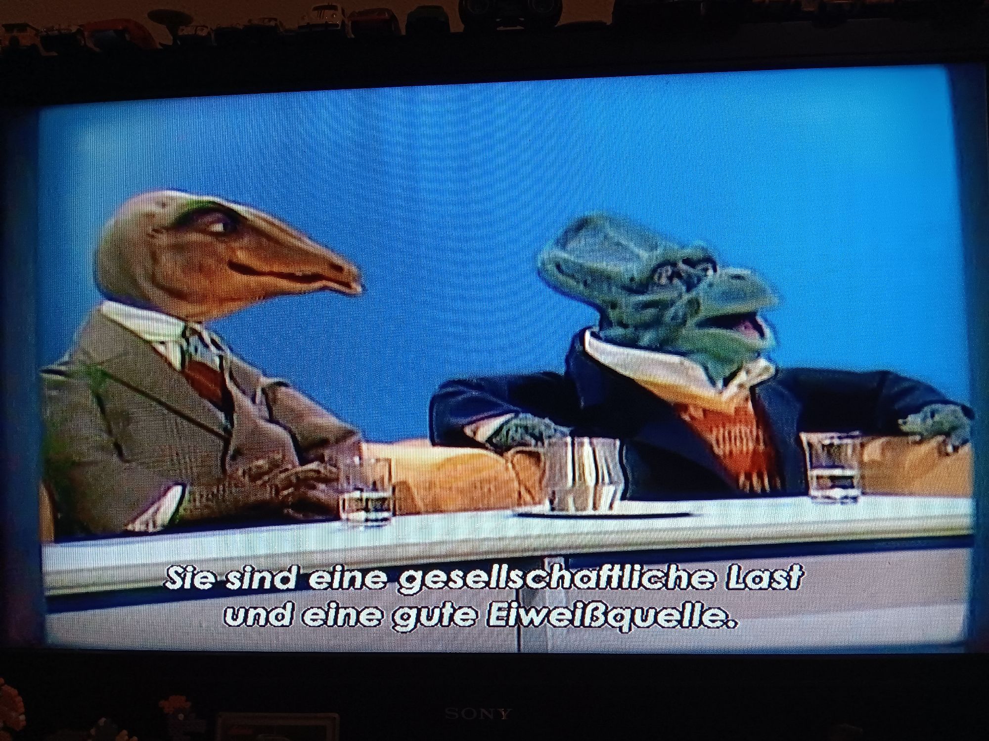 Szene aus der Puppensitcom DIE DINOS: zwei anthropomorphe Dinosauriermänner, ein Gallimimus und ein Protoceratops, die beide in scheinbar teure Klamotten gehüllt sind, sitzen nebeneinander. Der Protoceratopsmann sagt: "Sie sind eine gesellschaftliche Last und eine gute Eiweißquelle."