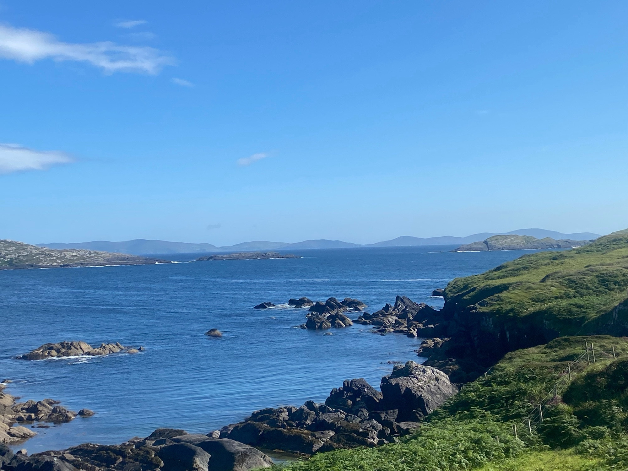 View across Derrynane Bay, County Kerry, over Kenmare Bay toward Beara Peninsula, County Cork on a sunny late afternoon in July, greenery and then rocks down to blue sea under a mostly clear blue sky toward the grey blue shadow of the distant Beara peninsula landscape.