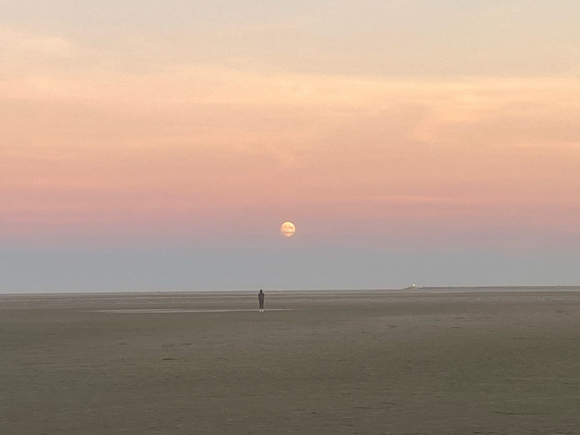 A woman stands in the middle distance of a beach at low tide just before sunset under a full moon in a pink yellow pale blue sky